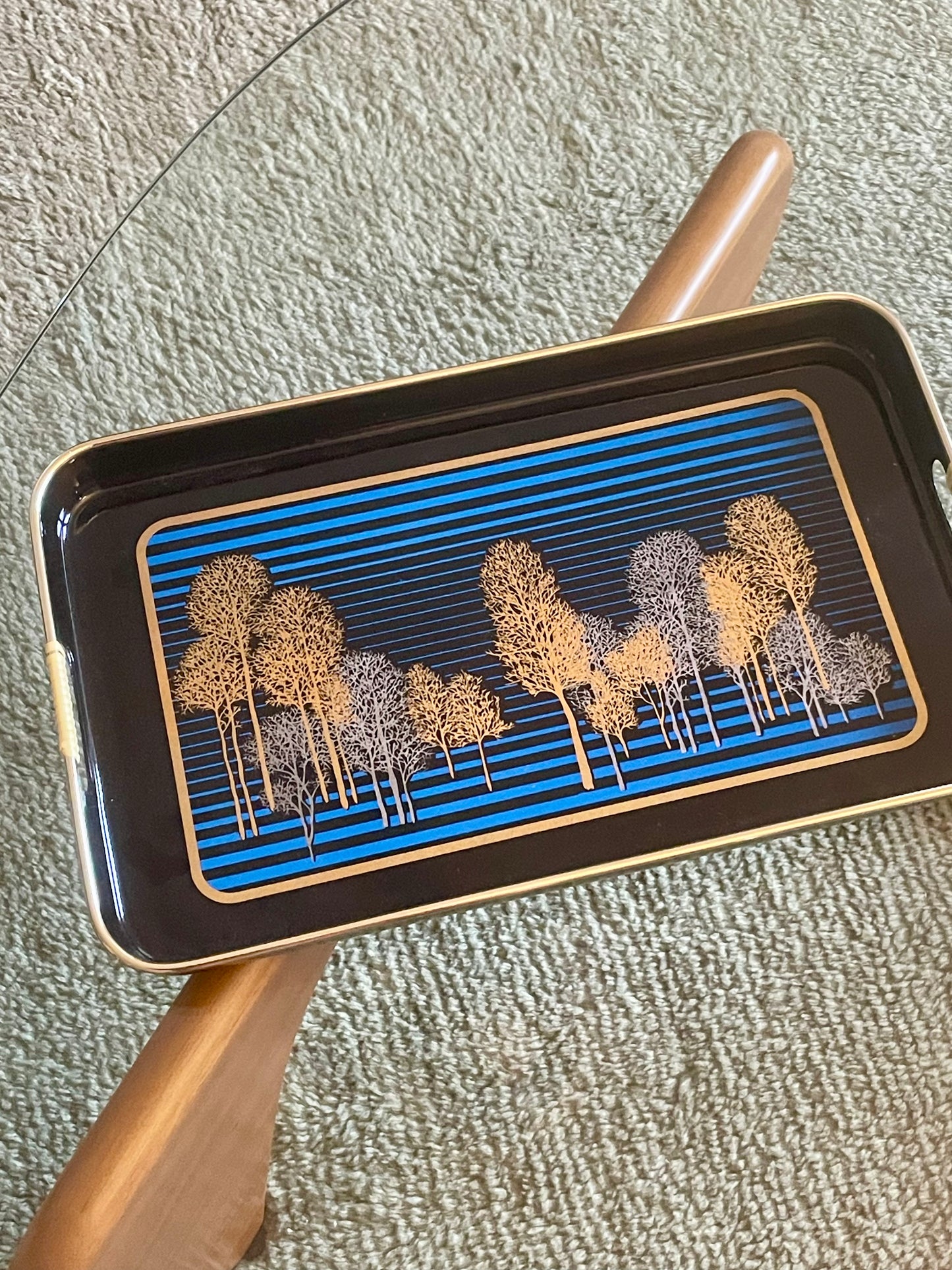 Rectangular black serving tray with gold-toned edges and handles, featuring a blue striped and gold and silver tree pattern, arranged on a glass table over a textured rug.