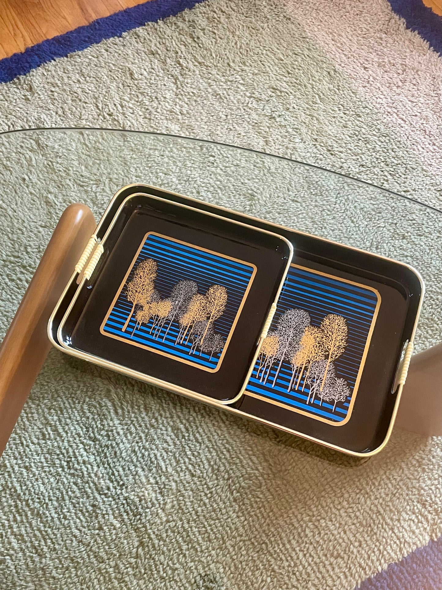 One rectangular and one square black serving tray with gold-toned edges and handles nested together, featuring a blue striped and gold and silver tree pattern, arranged on a glass table over a textured rug.
