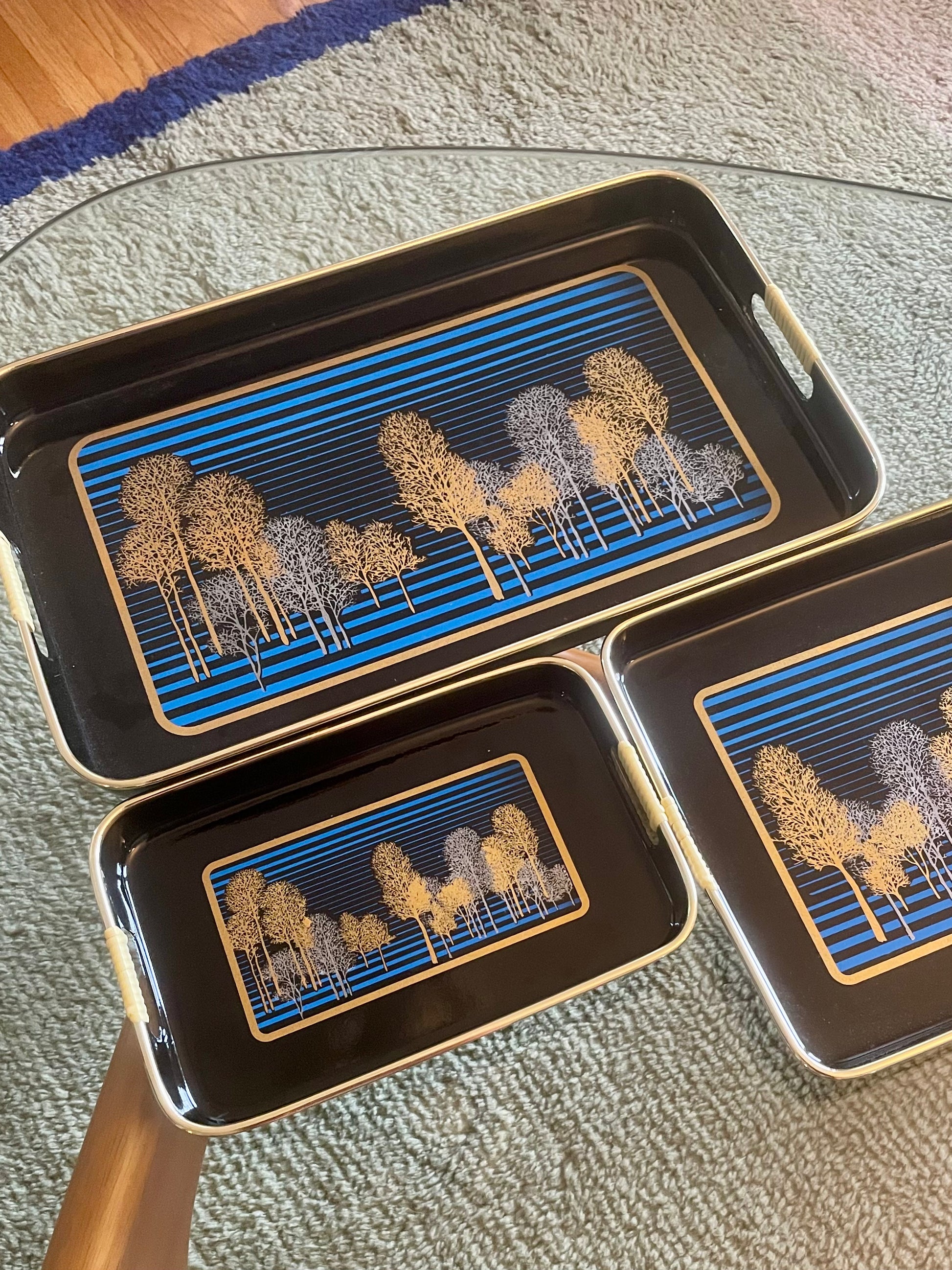 Three rectangular black serving trays with gold-toned edges and handles, featuring a blue striped and gold and silver tree pattern, arranged on a glass table over a textured rug.