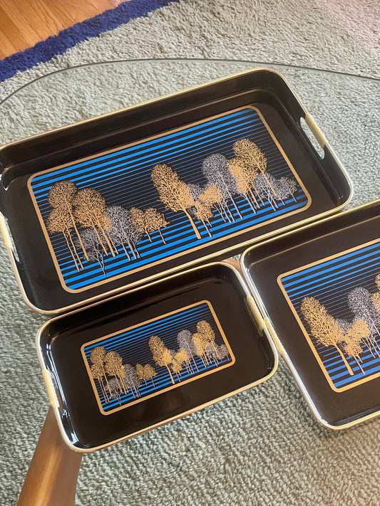 Three rectangular black serving trays with gold-toned edges and handles, featuring a blue striped and gold and silver tree pattern, arranged on a glass table over a textured rug.