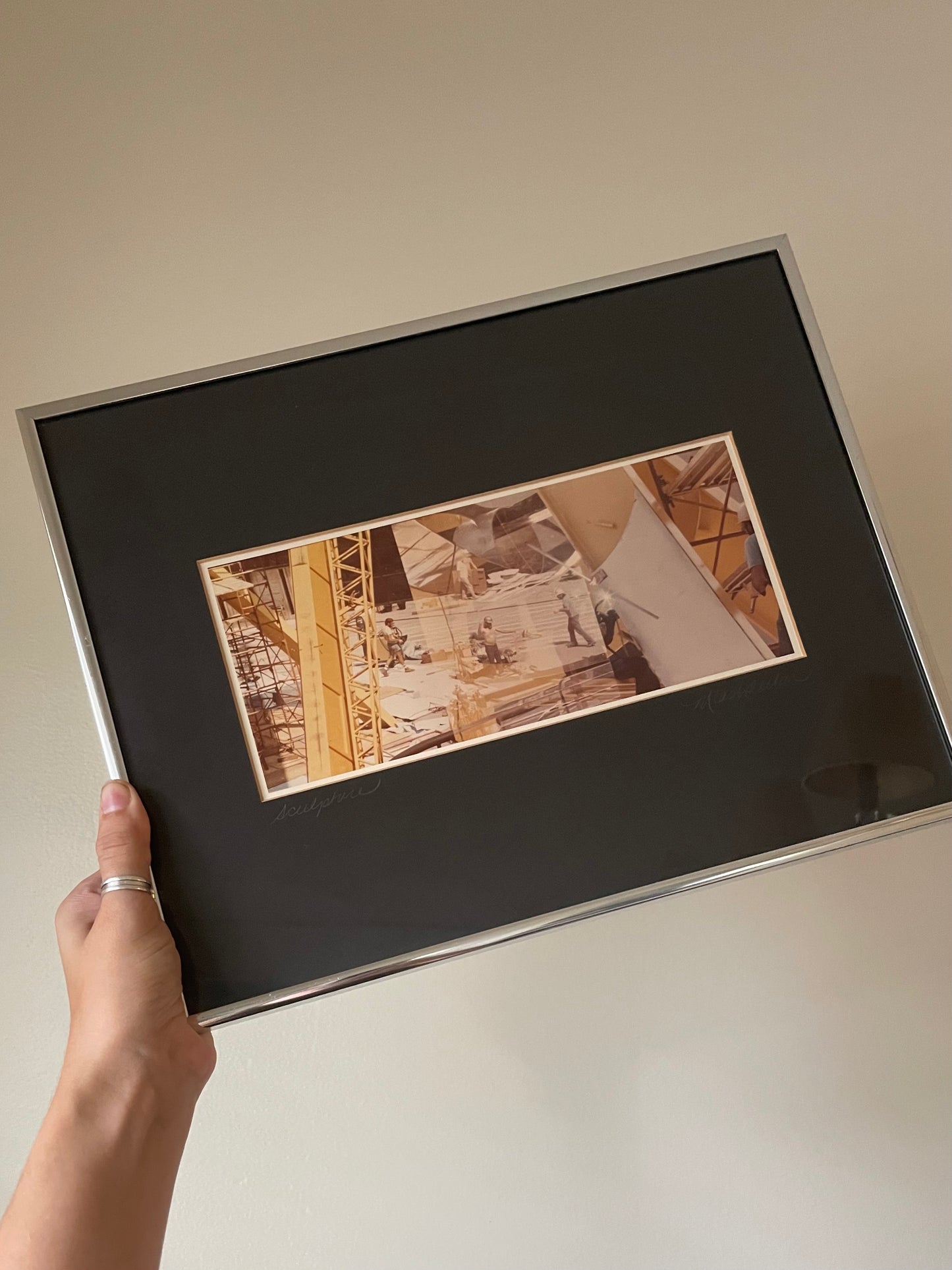 A hand holding a large framed art print with a black mat and a silver frame. The central print features a yellow and sepia-toned photograph of a construction site with cranes and workers.