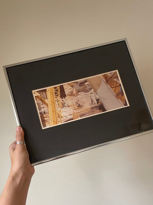 A hand holding a large framed art print with a black mat and a silver frame. The central print features a yellow and sepia-toned photograph of a construction site with cranes and workers.