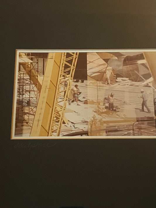 A framed yellow and sepia-toned photograph depicting construction workers on a building site with scaffolding and a crane, surrounded by a black mat and titled 'Sculpture' in handwritten script.