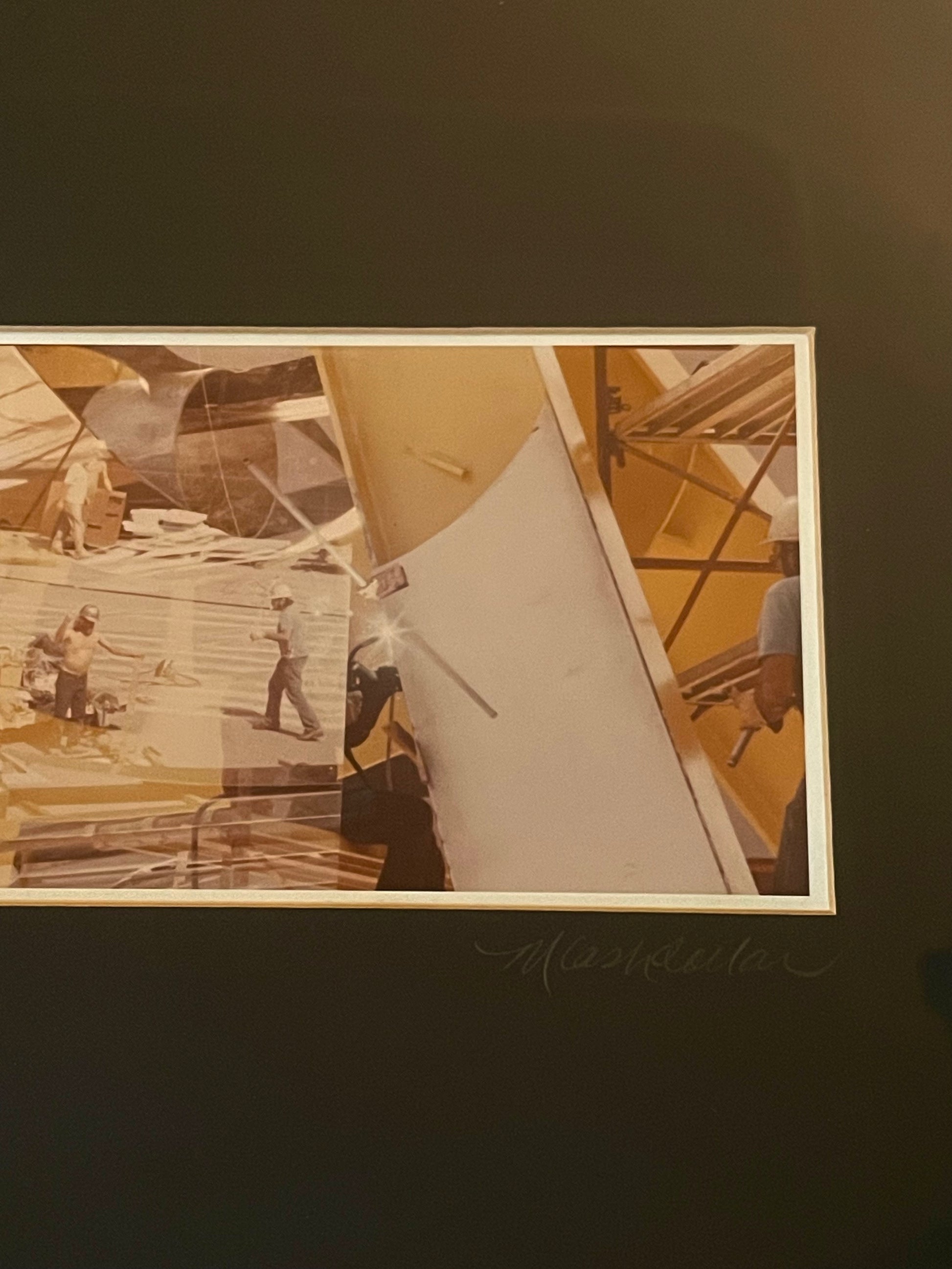 A framed yellow and sepia-toned photograph depicting construction workers on a building site with scaffolding and a crane, surrounded by a black mat and signed 'M. Cashdollar'.