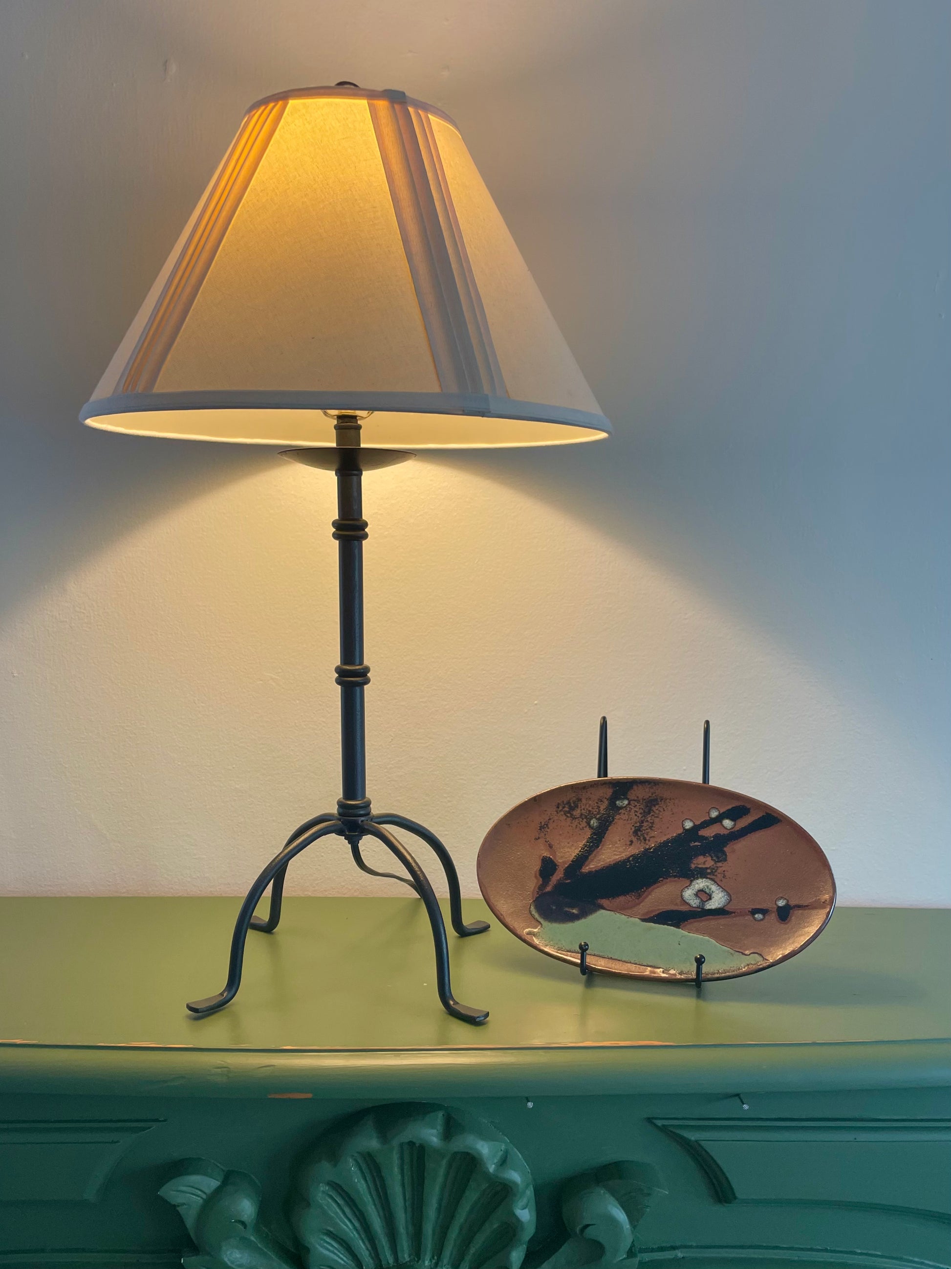 A table lamp with a warm glowing cream pleated shade and a black metal base with four penny feet, placed on a green mantel next to an oval-shaped ceramic platter with abstract glazing propped up in a dish stand.