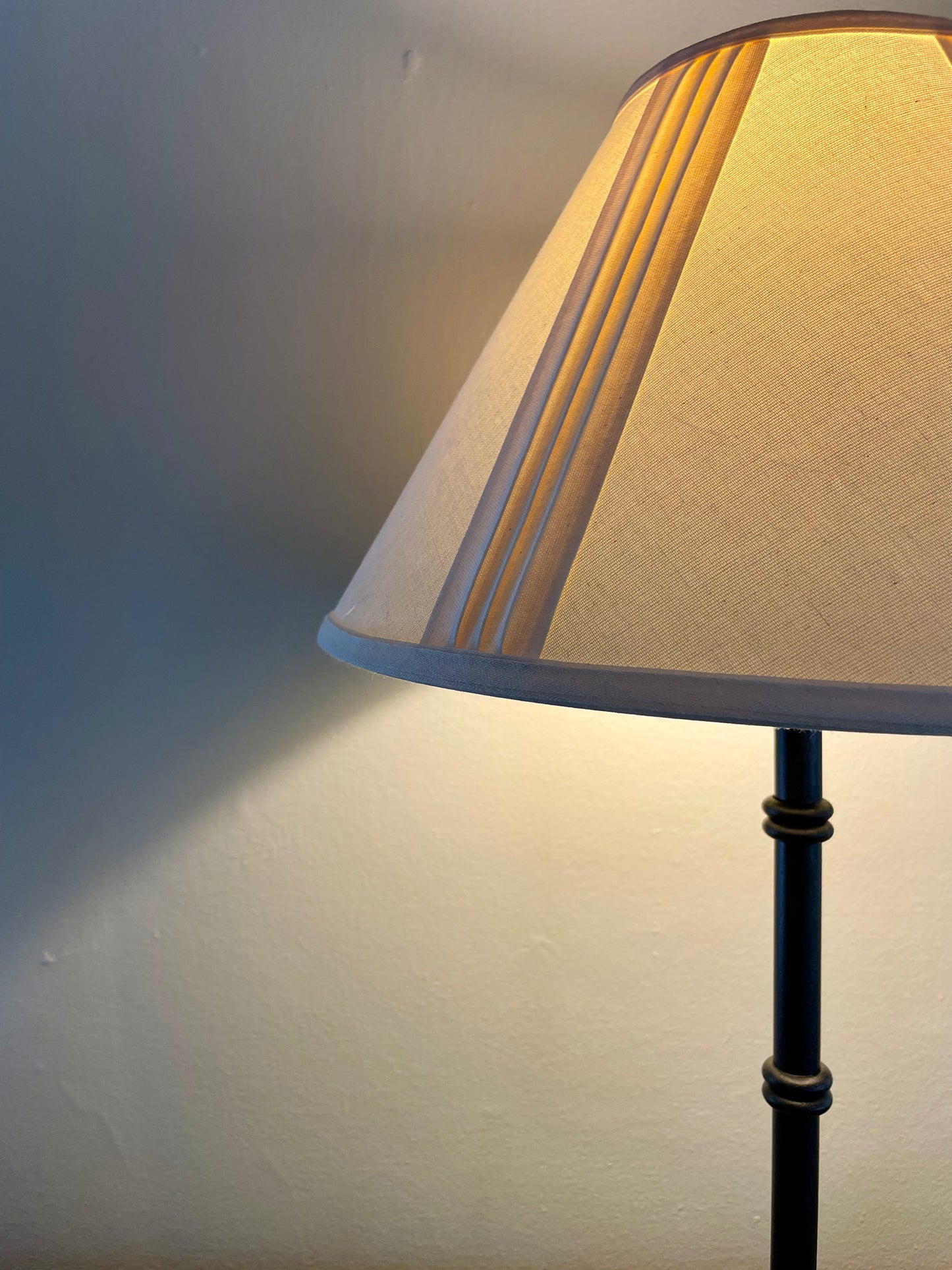 Closeup of a table lamp with a cream fabric shade and a black metal base with four penny feet, casting a warm yellow glow against a neutral wall.