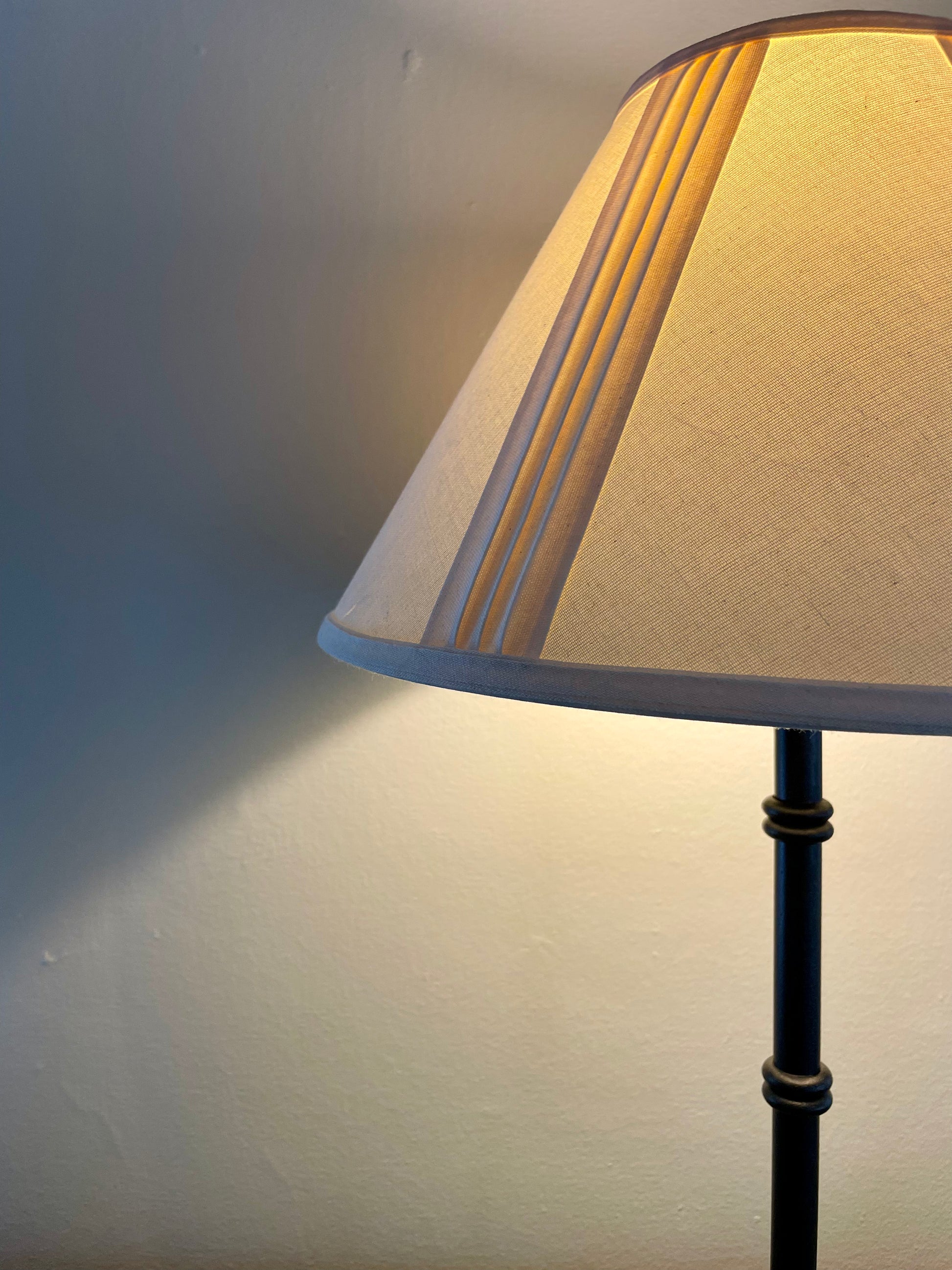 Closeup of a table lamp with a cream fabric shade and a black metal base with four penny feet, casting a warm yellow glow against a neutral wall.