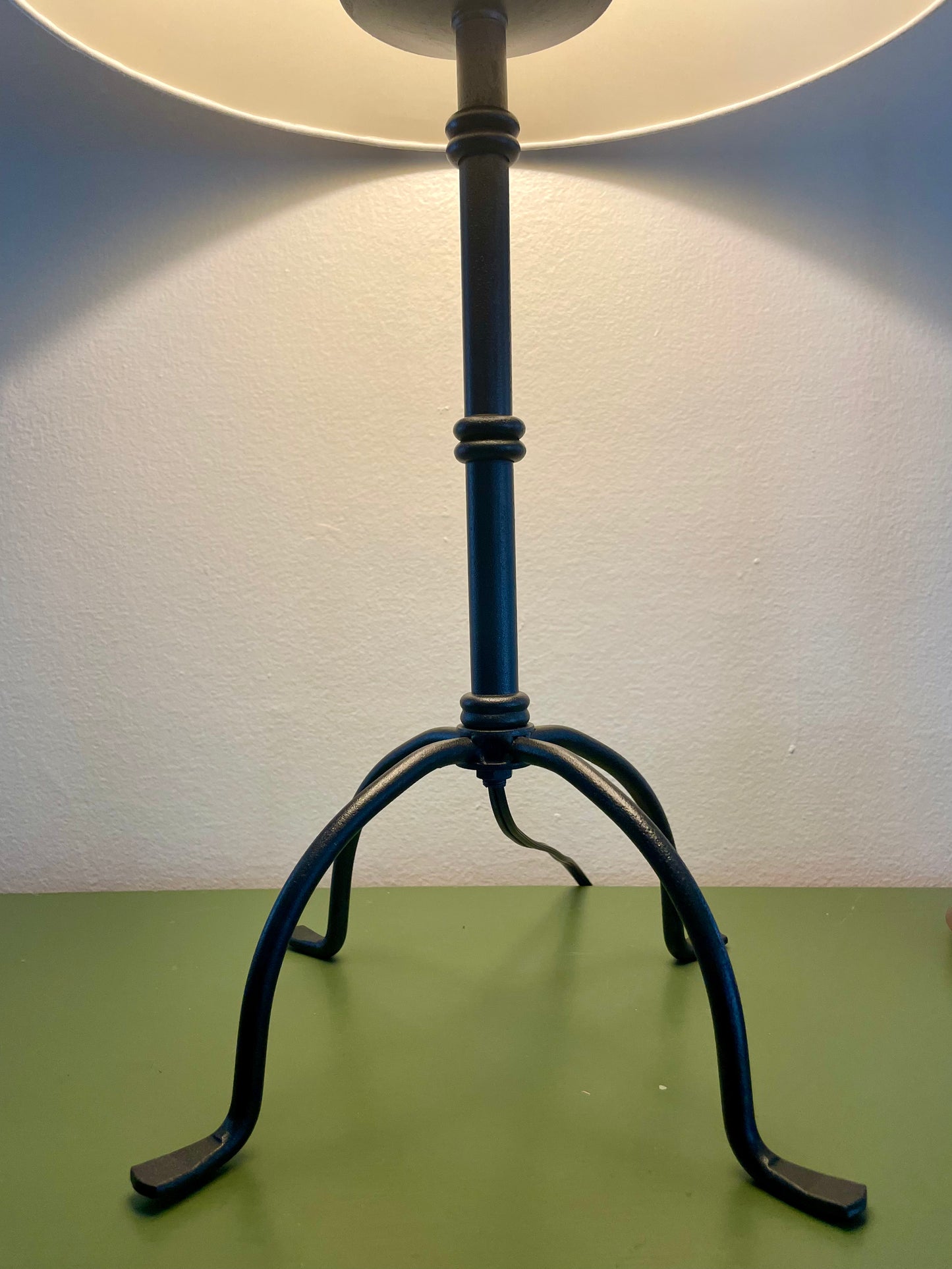 Closeup of a table lamp with a cream fabric shade and a black metal base with four penny feet, shown lit against a plain wall and green tabletop.