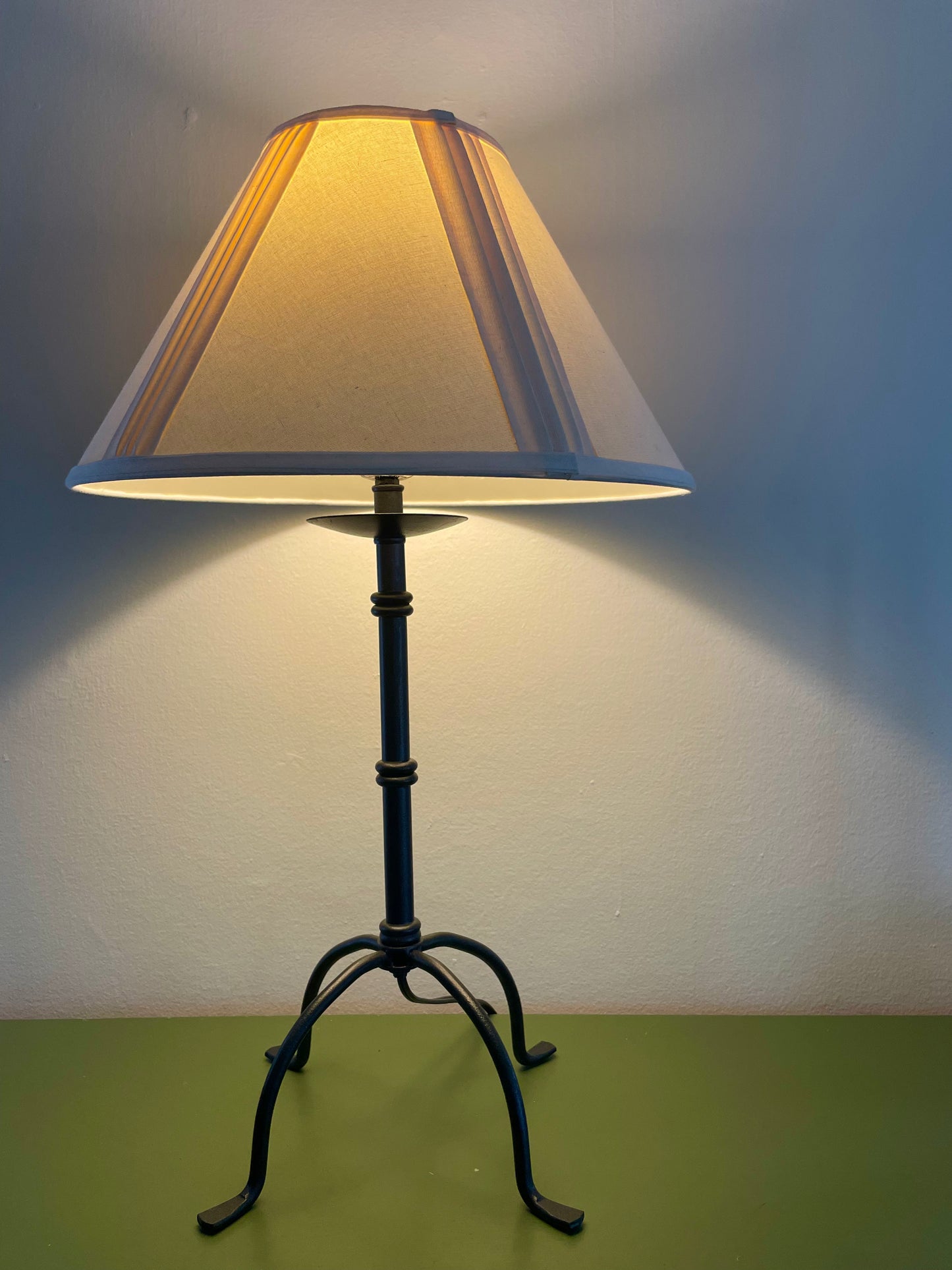 A table lamp with a cream fabric shade and a black metal base with four penny feet, shown lit against a plain wall and green tabletop.