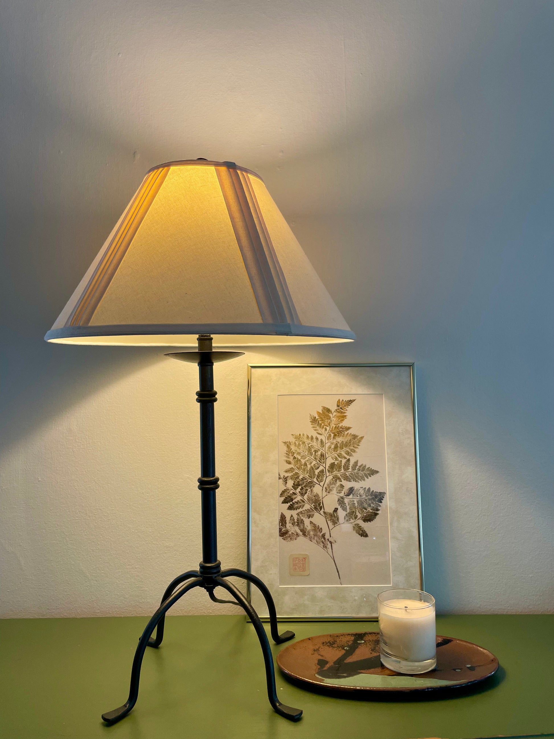 A table lamp with a black metal base with four penny feet and a cream pleated fabric shade, glowing warm yellow. It is styled on a green surface next to a framed botanical print and a lit candle on a ceramic dish, against a blue-gray wall.