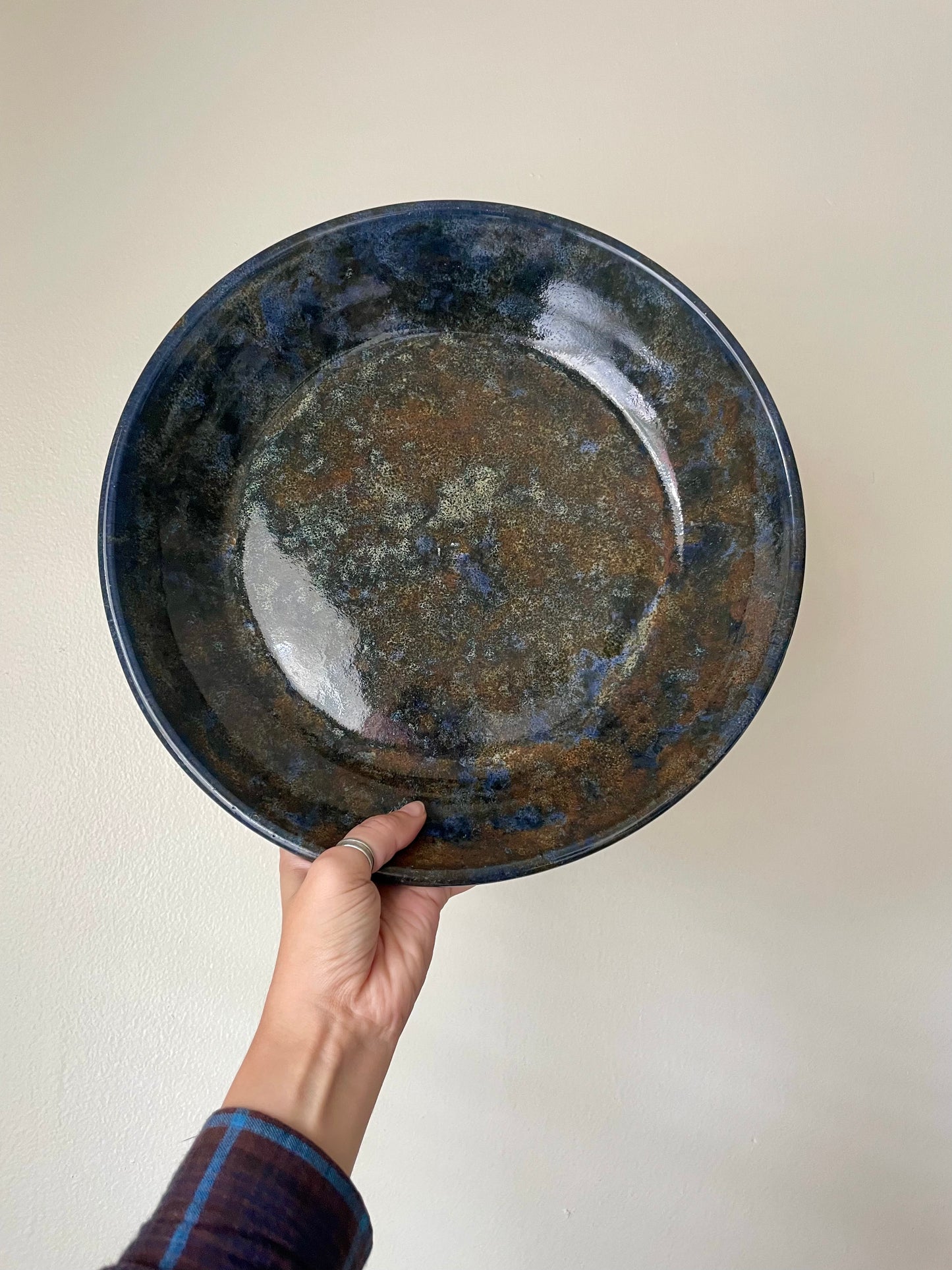 A hand holds a large round ceramic bowl with speckled blue and brown glazing in front of a plain wall.