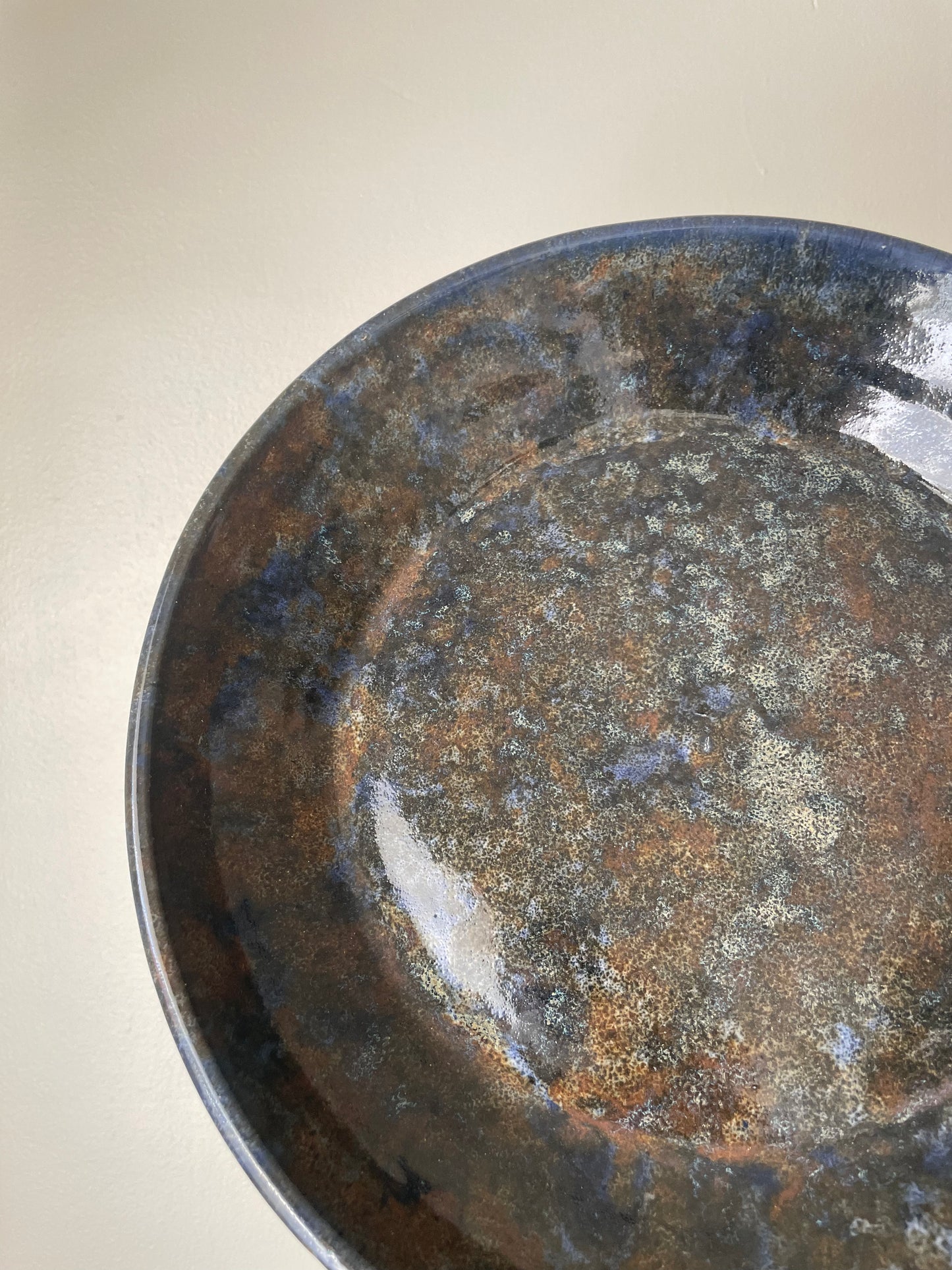 Close-up of a ceramic plate with a mottled glaze in dark blue, brown, and beige tones, photographed against a plain light background.