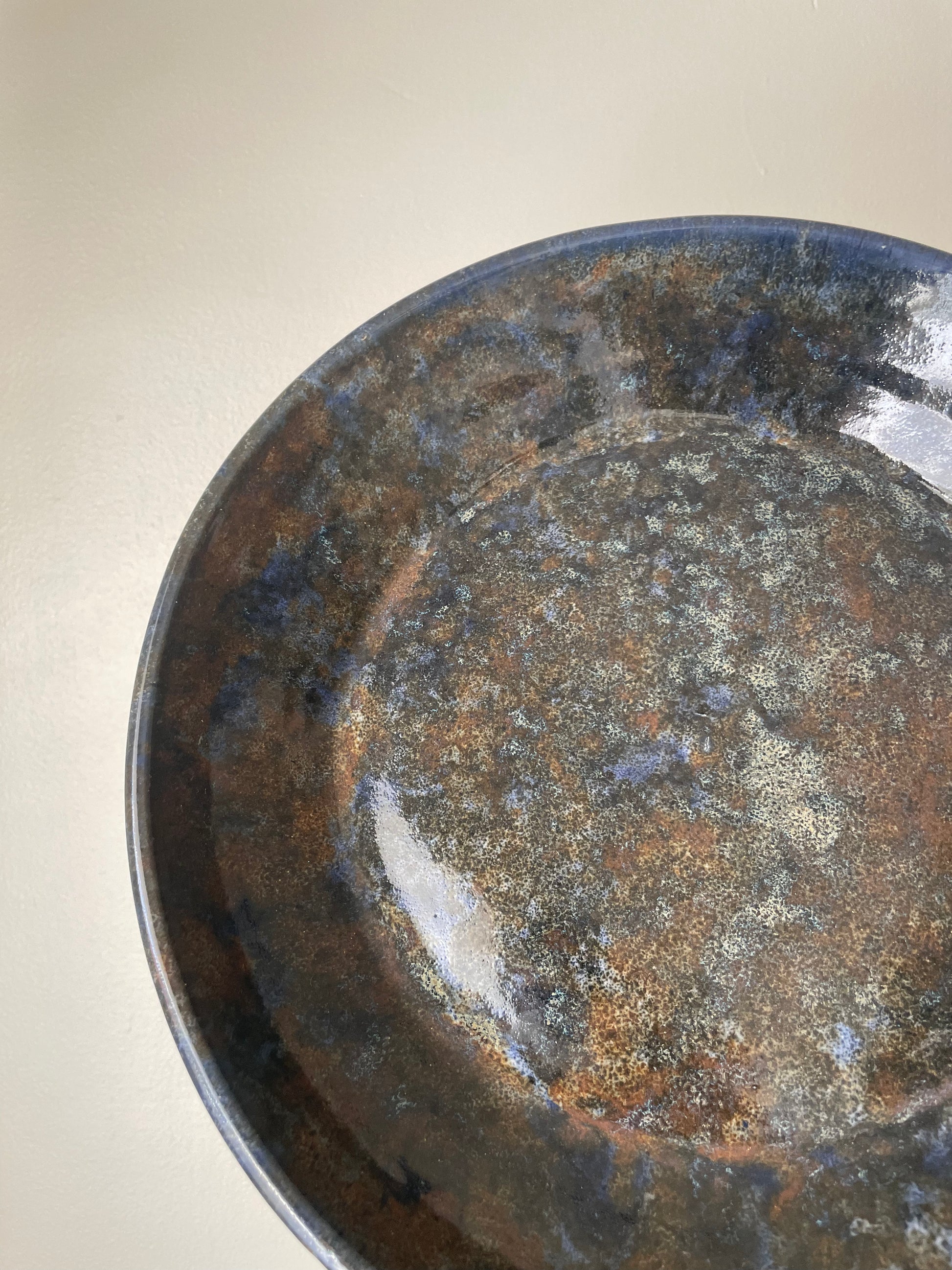 Close-up of a ceramic plate with a mottled glaze in dark blue, brown, and beige tones, photographed against a plain light background.