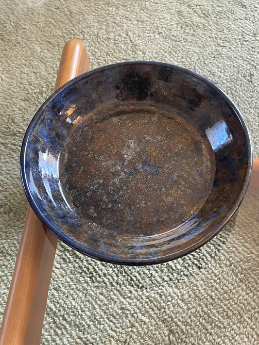 A large round ceramic bowl with speckled blue and brown glazing rests on a glass table over a blue-grey rug.