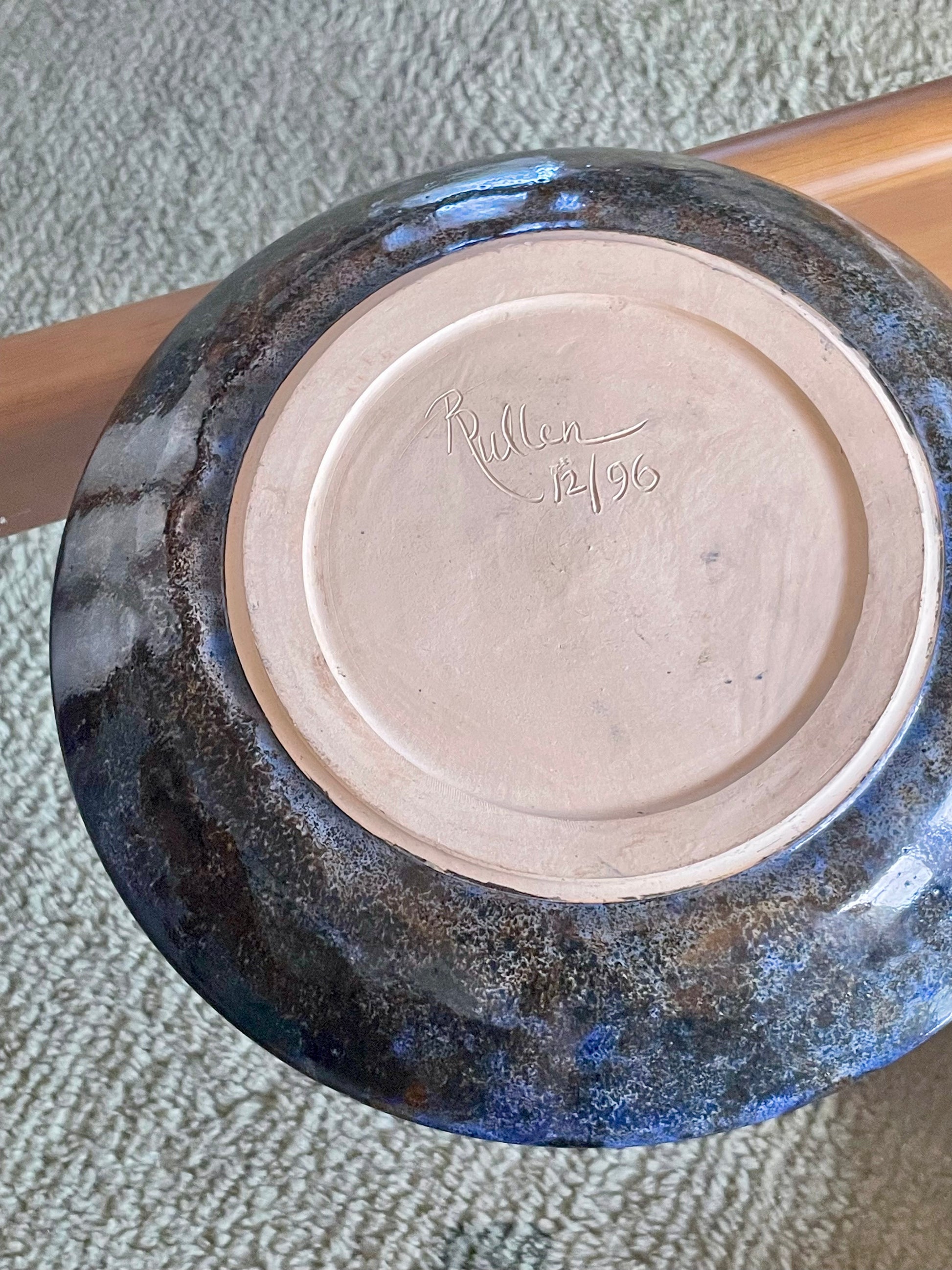 A large ceramic bowl rests upside down on a glass table over a blue-grey rug, showing a signature reading "R Pullen 12/96".