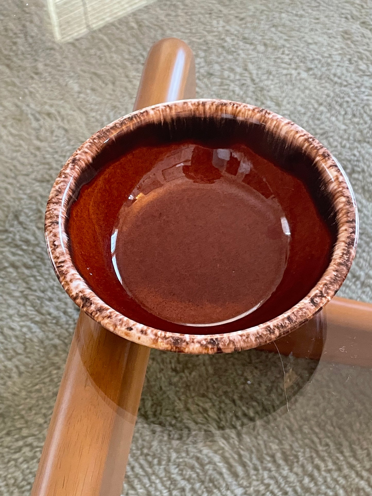 A handmade ceramic bowl with a glossy reddish-brown interior and a textured, speckled brown and beige exterior, shown resting on a glass-top wooden table.