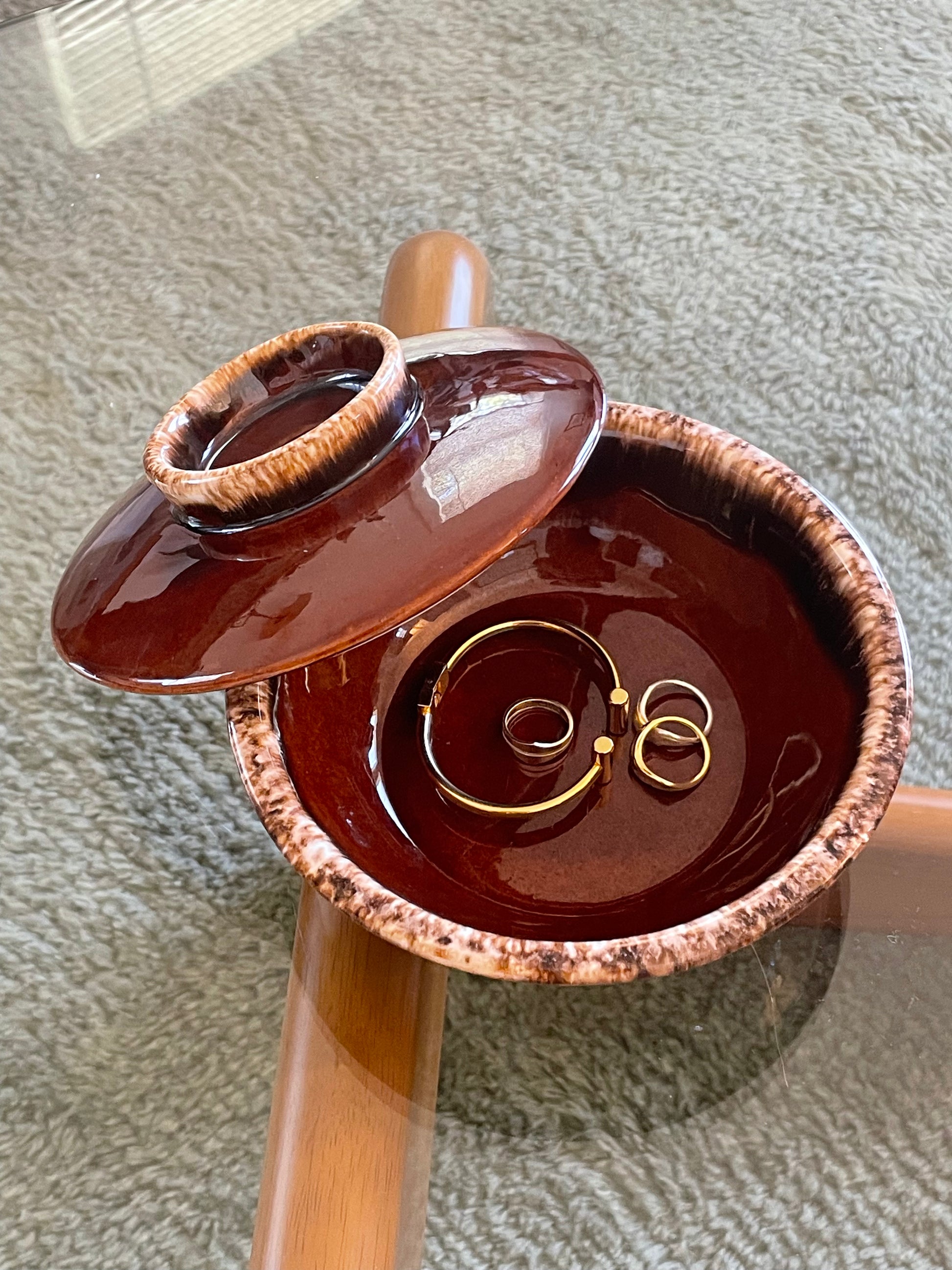A glossy dark brown ceramic jewelry dish with a matching lid, shown open on a glass-top wooden table over a gray textured carpet. Inside the box are a bracelet and three rings.