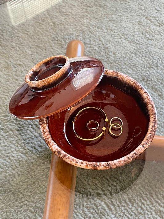 A glossy dark brown ceramic jewelry dish with a matching lid, shown open on a glass-top wooden table over a gray textured carpet. Inside the box are a bracelet and three rings.