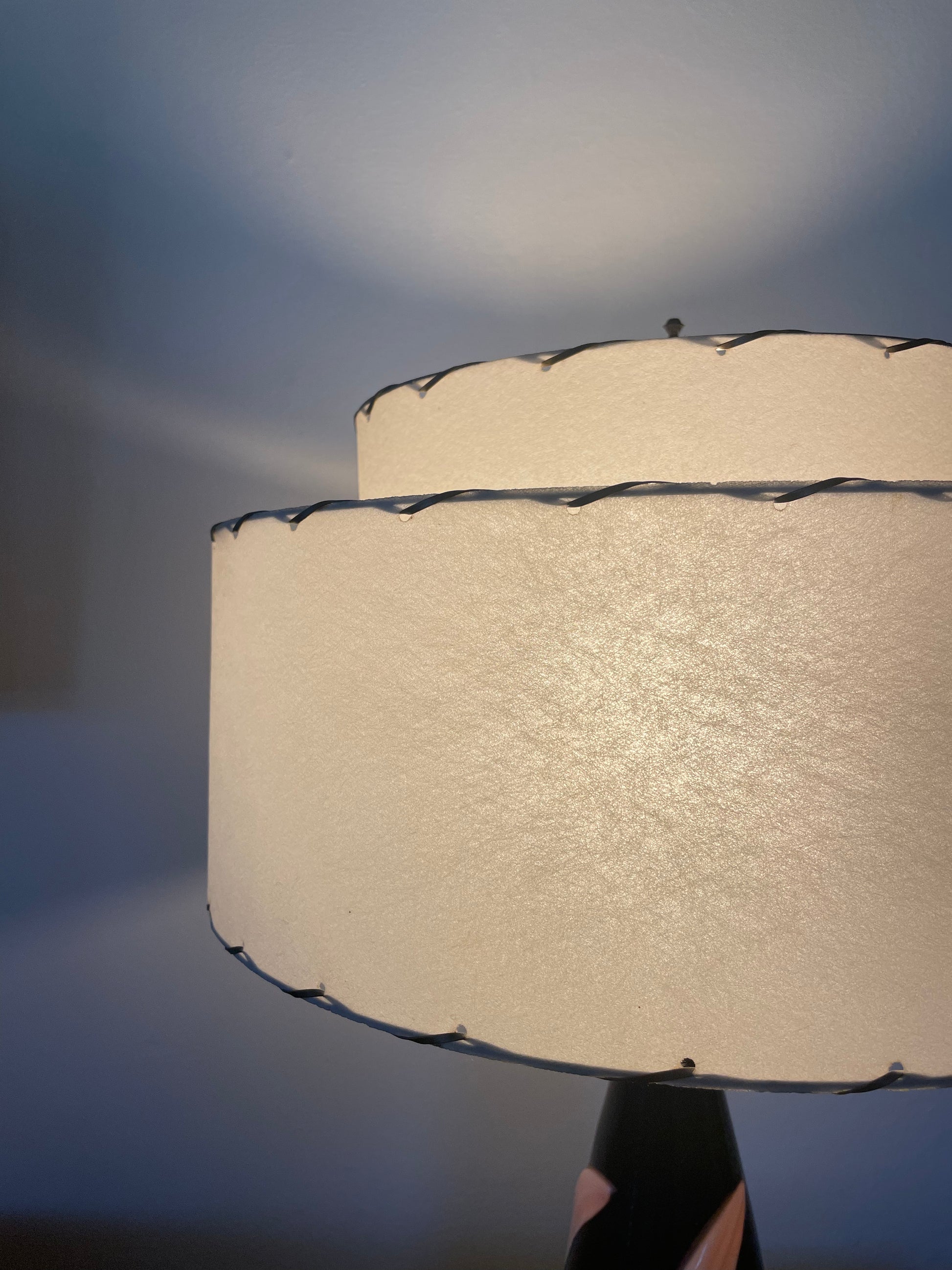 A close-up of a lit table lamp with two stacked cylindrical fiberglass shades, finished with dark laced edges, set against a dark grey background.