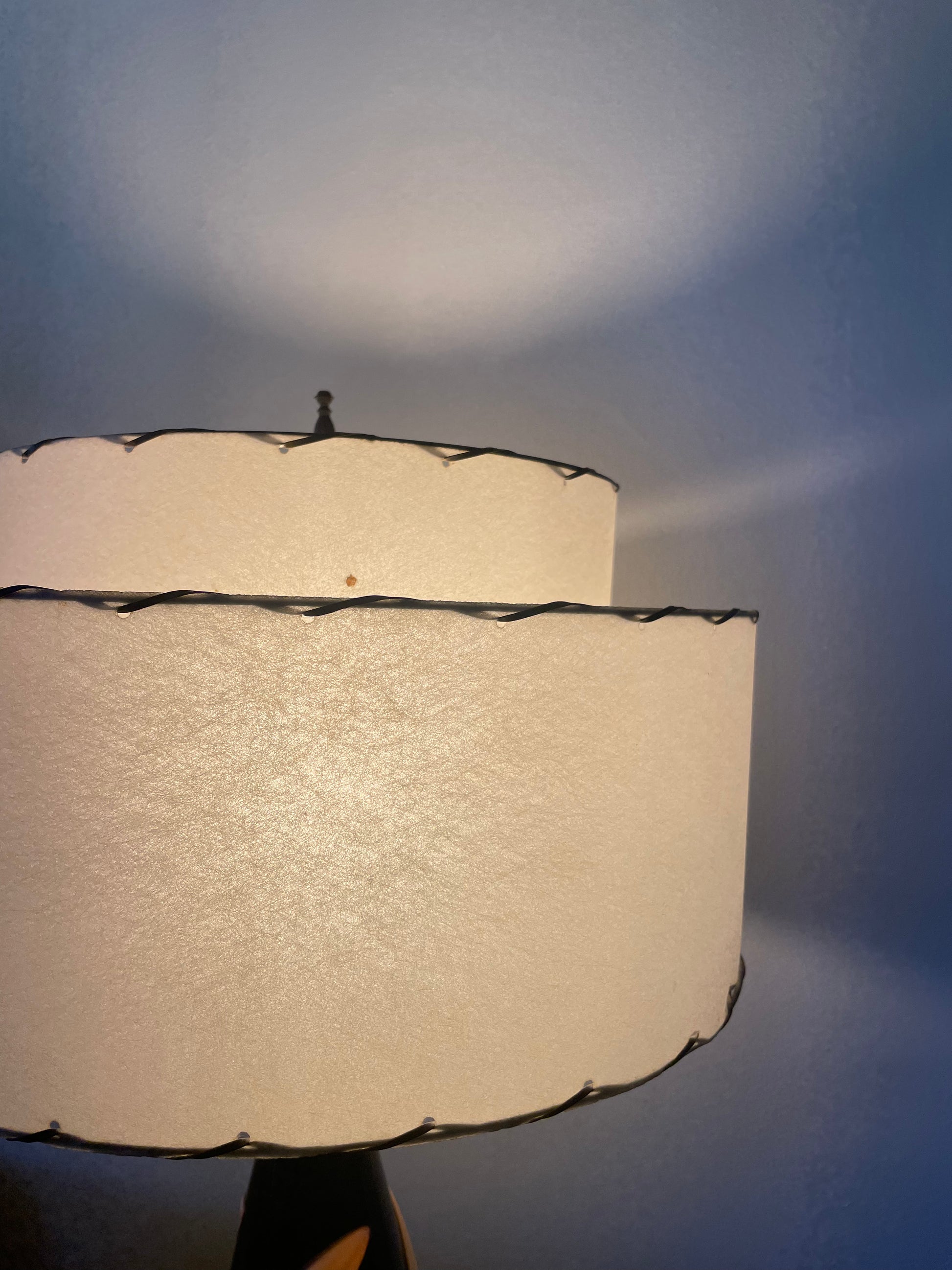 A close-up of a lit table lamp with two stacked cylindrical fiberglass shades, finished with dark laced edges, set against a dark grey background.