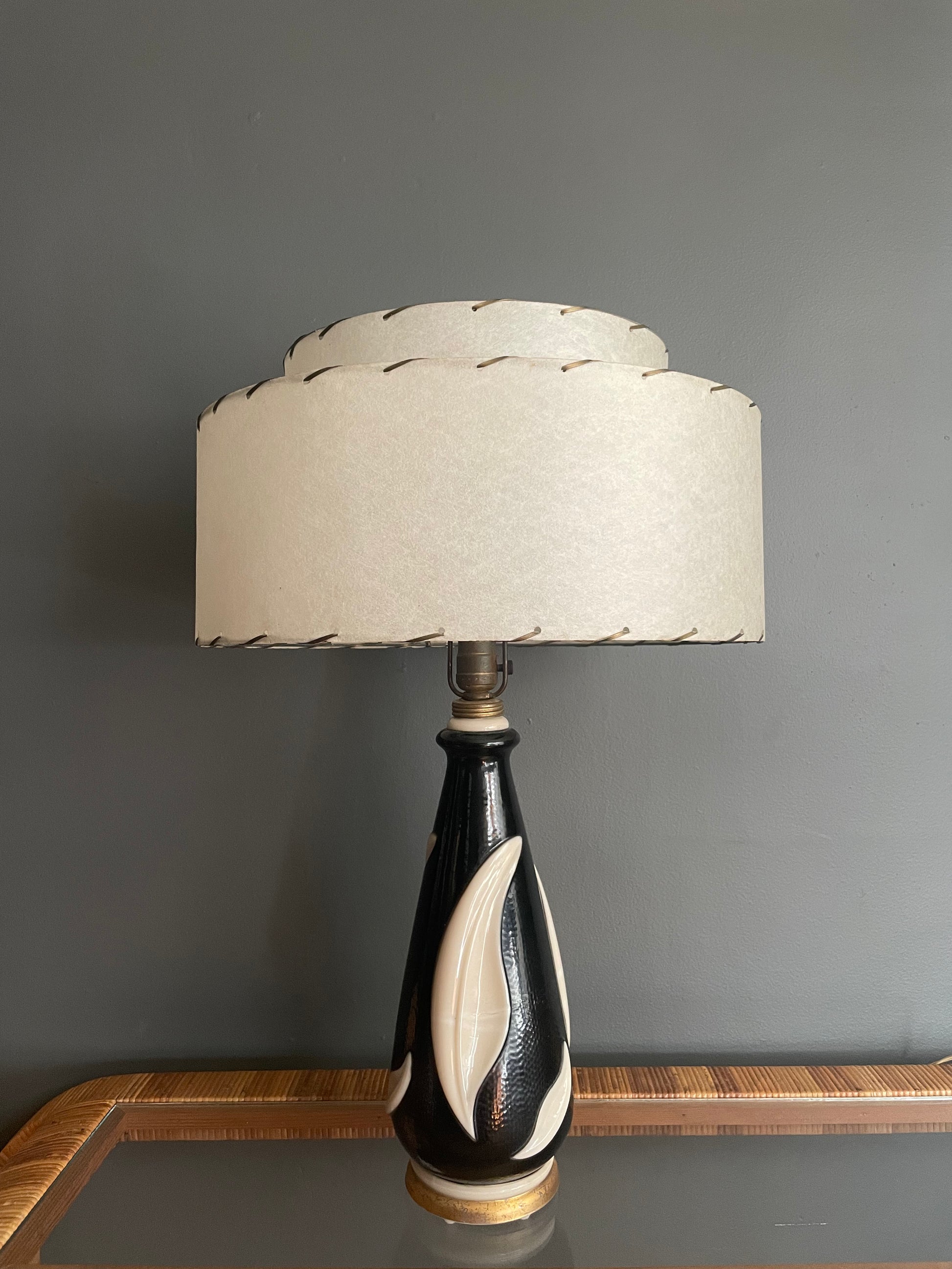 A table lamp with a black and cream sculptural glass base and a tiered cream fiberglass drum shade, set  against a dark gray wall.
