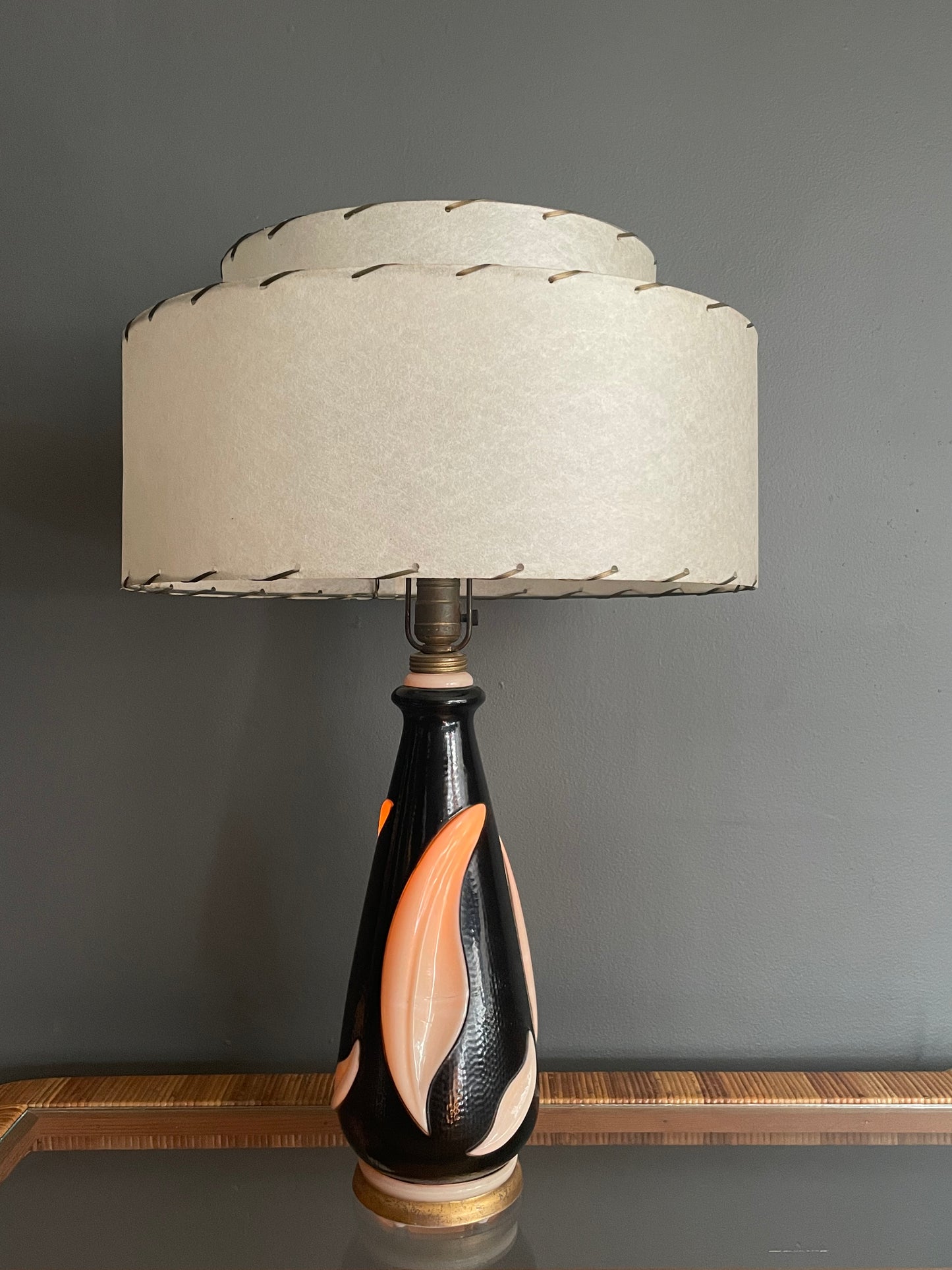 A table lamp with a tiered cream fiberglass shade and a glossy black ceramic base featuring a cream leaf motif, illuminated from within for a warm orange glow and set against a dark gray wall.