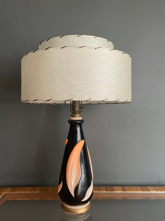 A table lamp with a tiered cream fiberglass shade and a glossy black ceramic base featuring a cream leaf motif, illuminated from within for a warm orange glow and set against a dark gray wall.