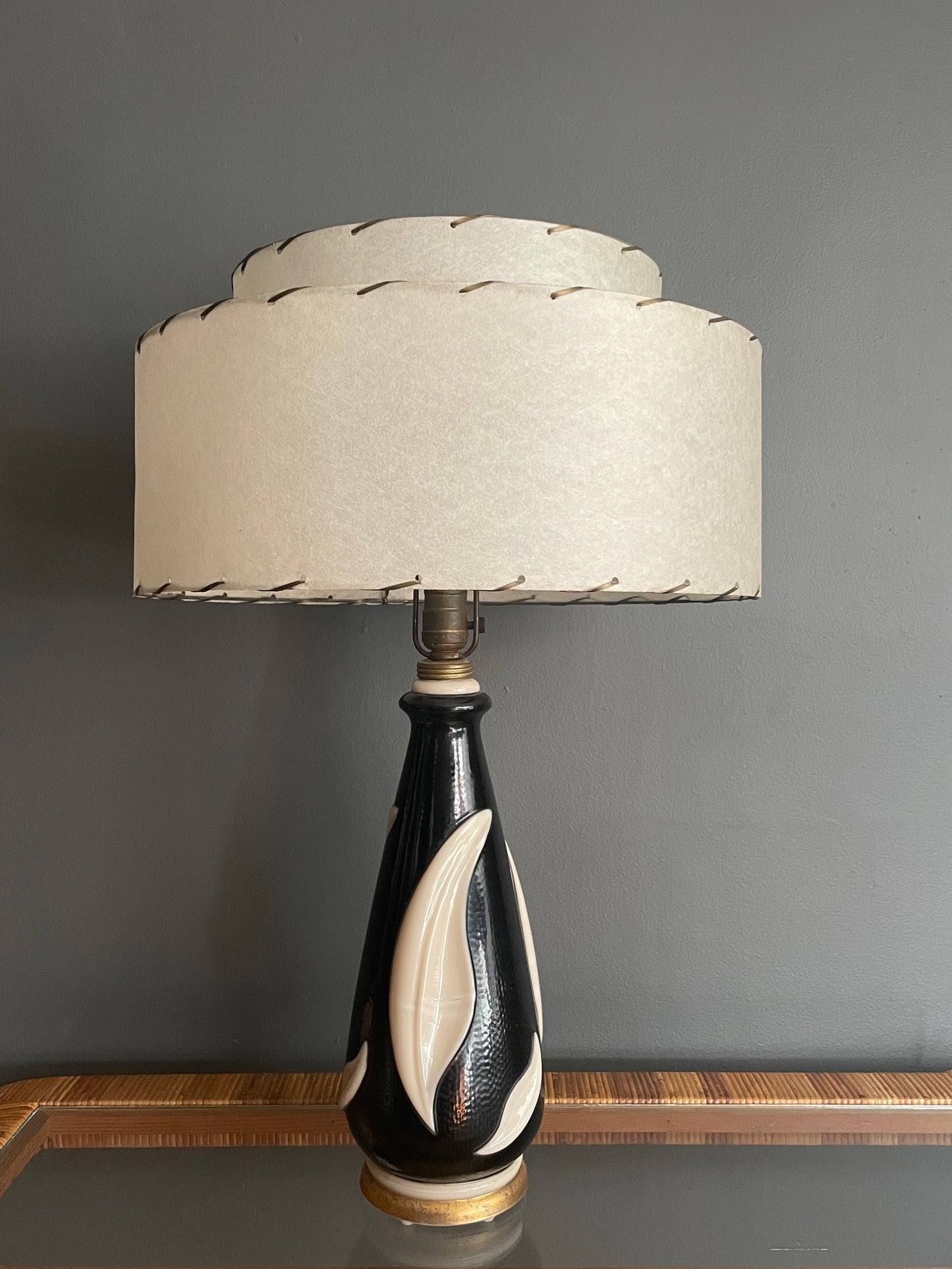 A table lamp with a tiered cream fiberglass shade and a glossy black glass base featuring sculpted cream leaf forms, set against a dark gray wall.