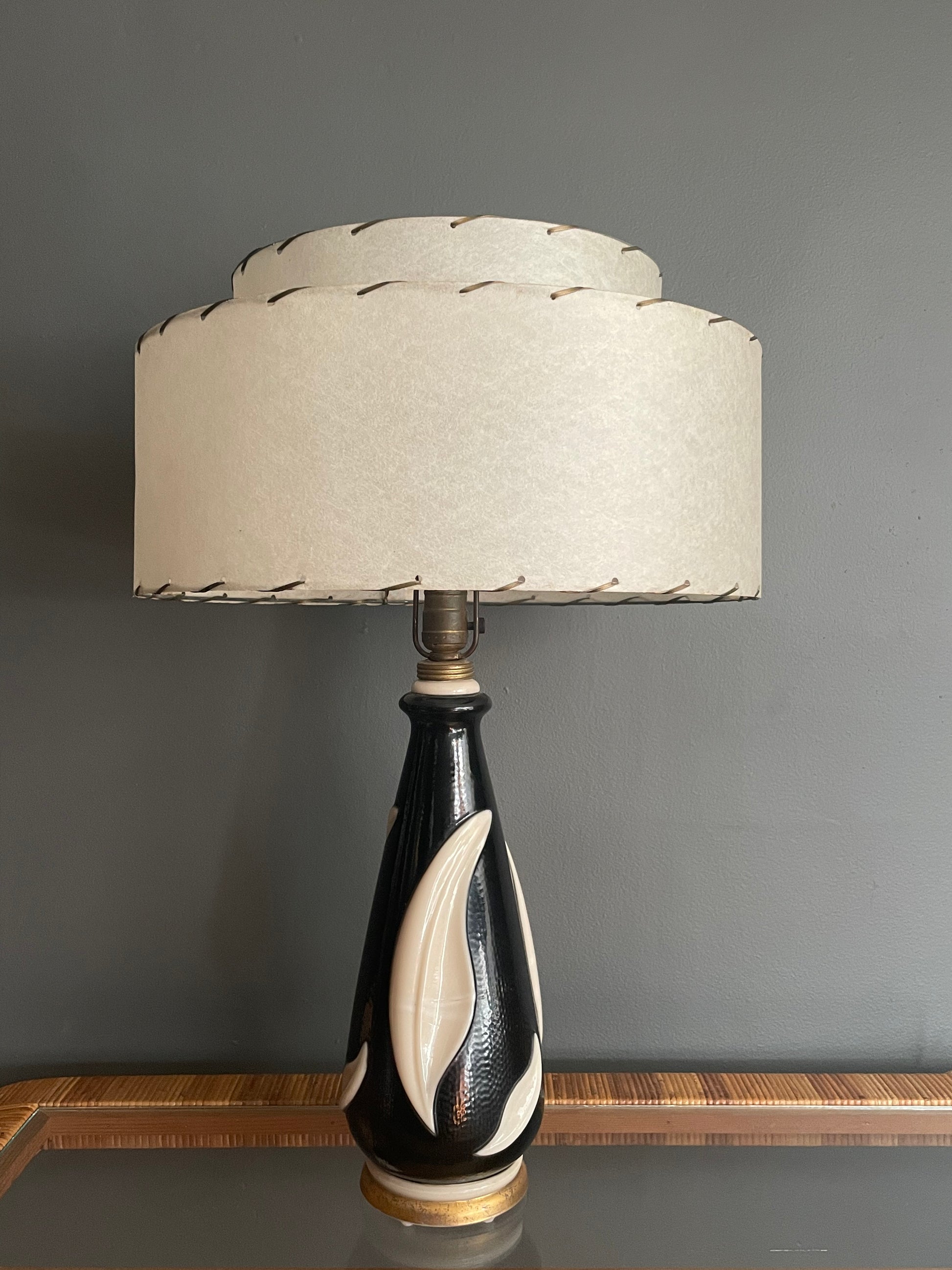 A table lamp with a tiered cream fiberglass shade and a glossy black glass base featuring sculpted cream leaf forms, set against a dark gray wall.