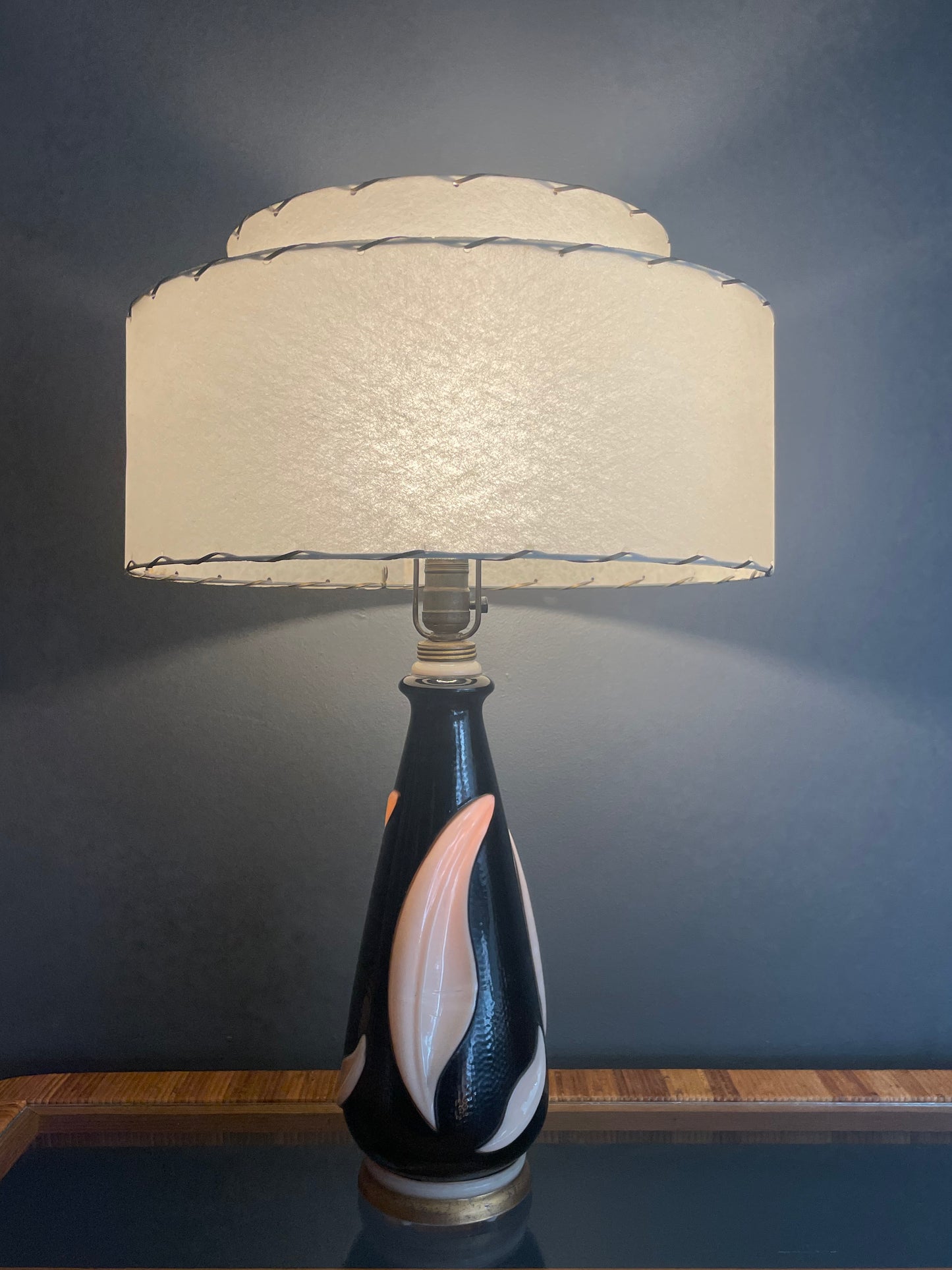 A table lamp with a textured cream-colored tiered drum shade and a glossy black painted glass base featuring a large cream leaf accent, illuminated by the bulb and from within, set against a dark gray wall.