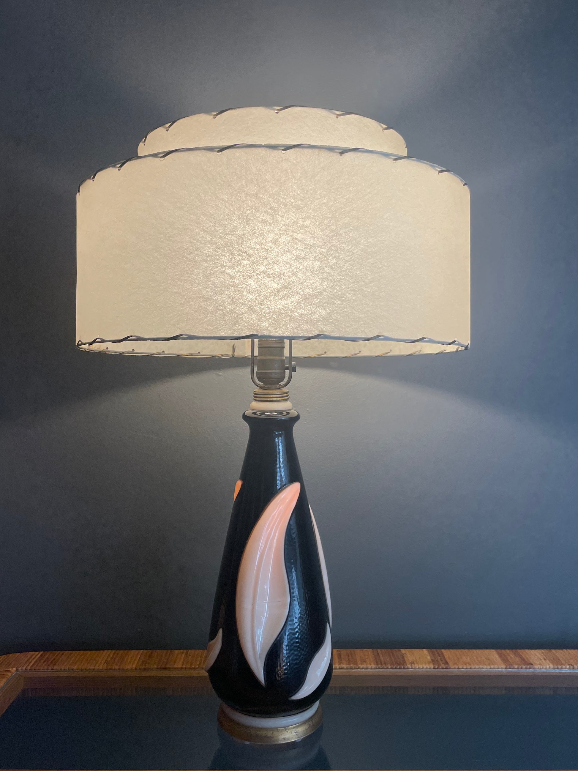 A table lamp with a textured cream-colored tiered drum shade and a glossy black painted glass base featuring a large cream leaf accent, illuminated by the bulb and from within, set against a dark gray wall.