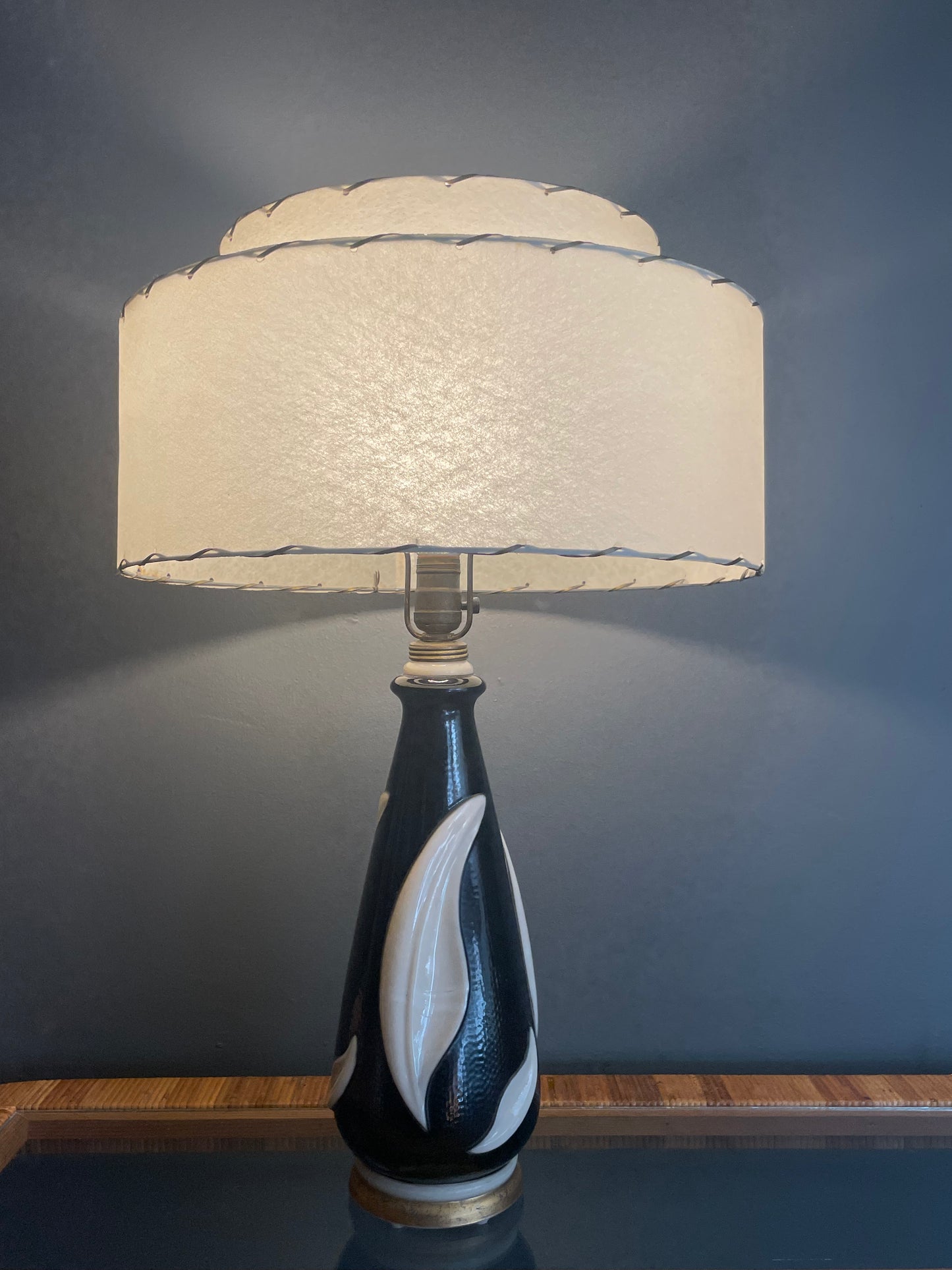 A table lamp with a cream-colored tiered fiberglass drum shade and a glossy black painted glass base featuring white leaf-like patterns, set against a dark gray wall.