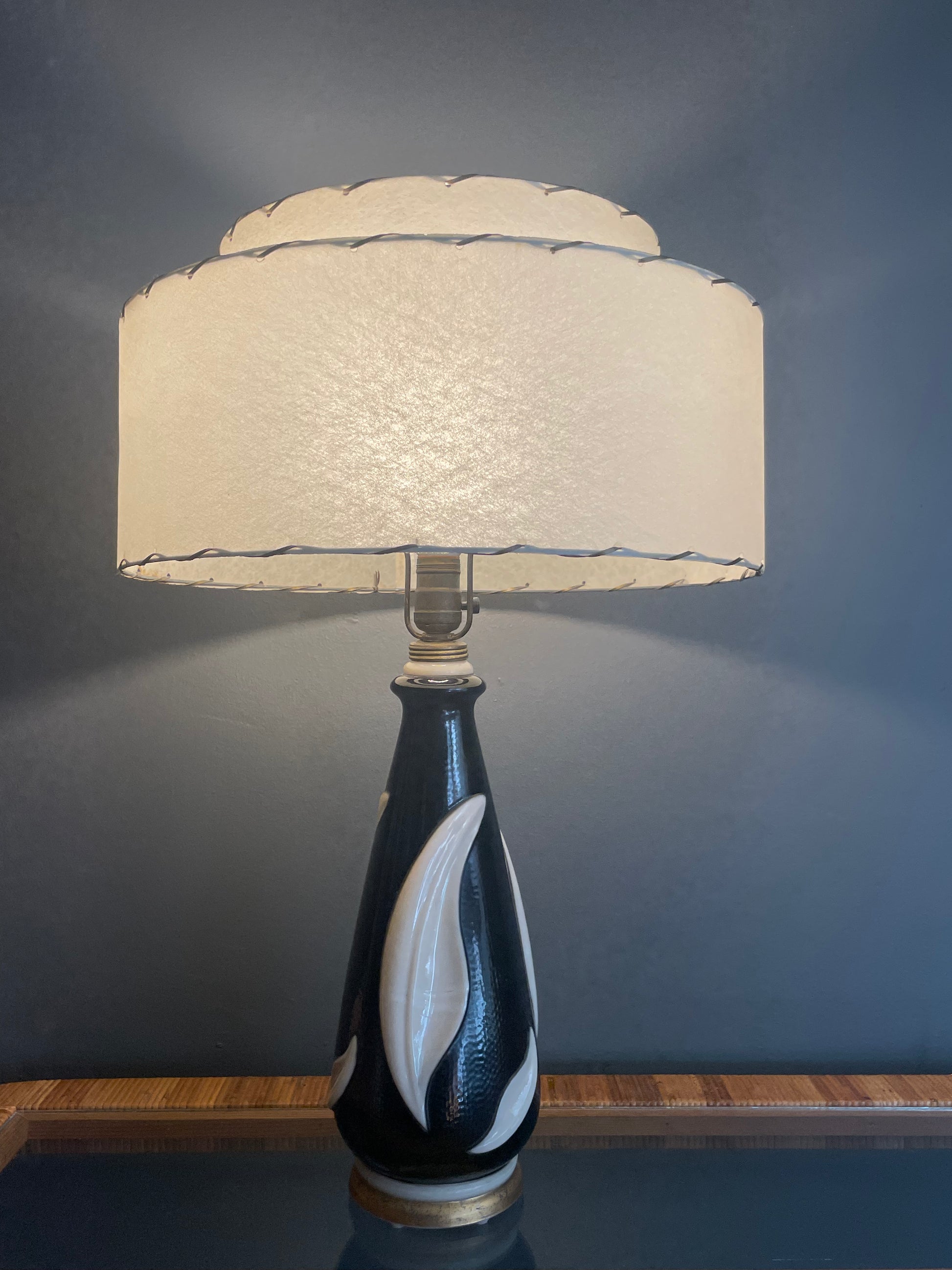 A table lamp with a cream-colored tiered fiberglass drum shade and a glossy black painted glass base featuring white leaf-like patterns, set against a dark gray wall.