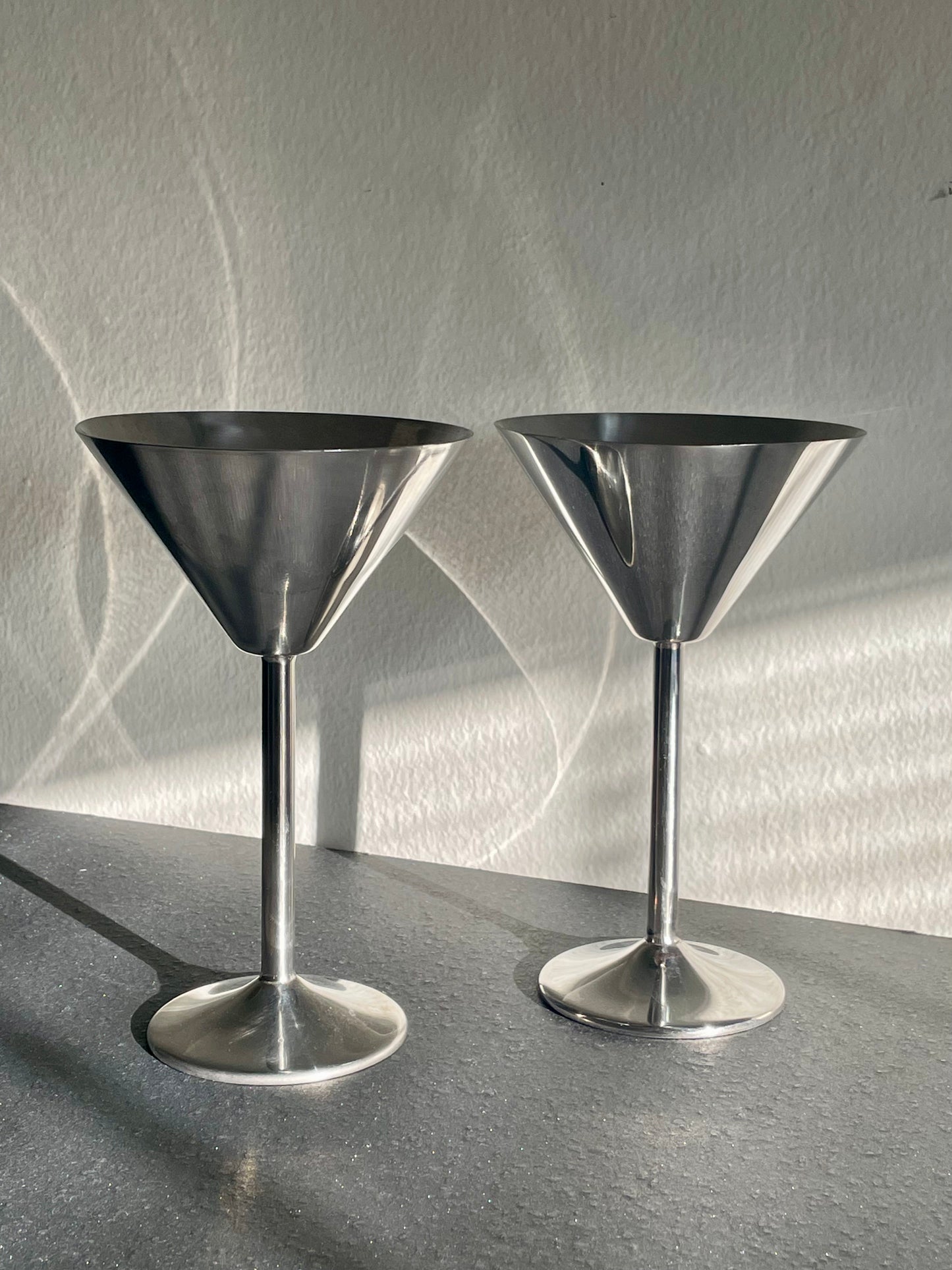 Two polished silver metal cocktail muddlers with wide conical heads and slender stems, standing on a dark gray surface against a textured light gray wall with dramatic shadows.