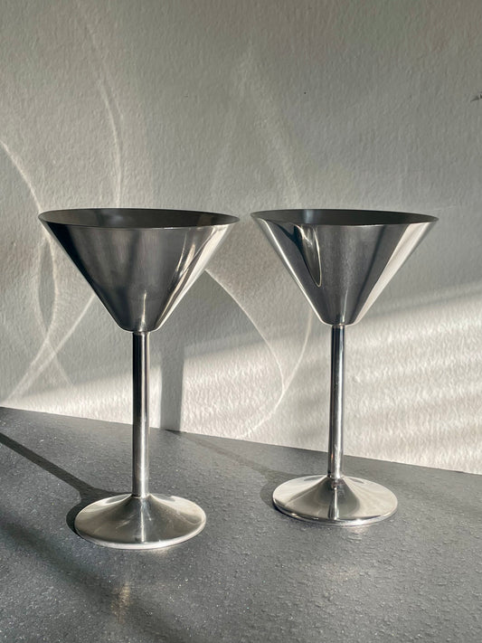 Two polished silver metal cocktail muddlers with wide conical heads and slender stems, standing on a dark gray surface against a textured light gray wall with dramatic shadows.