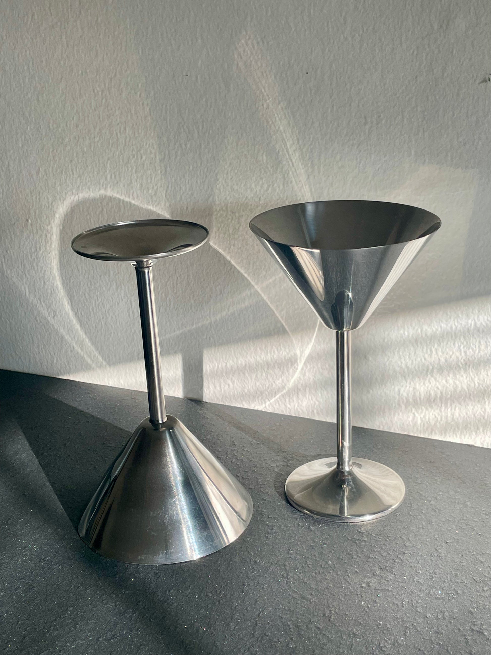 Two modern stainless steel martini glasses with conical metal bases, shown with one standing upright and one upside down on a dark tabletop against a textured light wall with strong natural shadows.