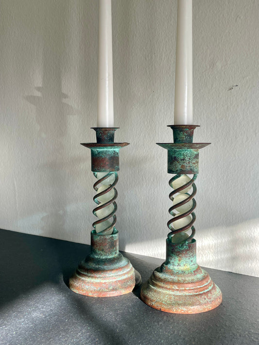 A pair of vintage-style metal candlestick holders with twisted spiral centers, showing a green and brown patina finish, holding white taper candles against a light wall.