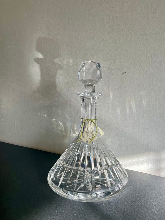 A clear cut-crystal decanter with a matching faceted stopper, resting on a dark surface against a light wall with a soft shadow cast behind it.