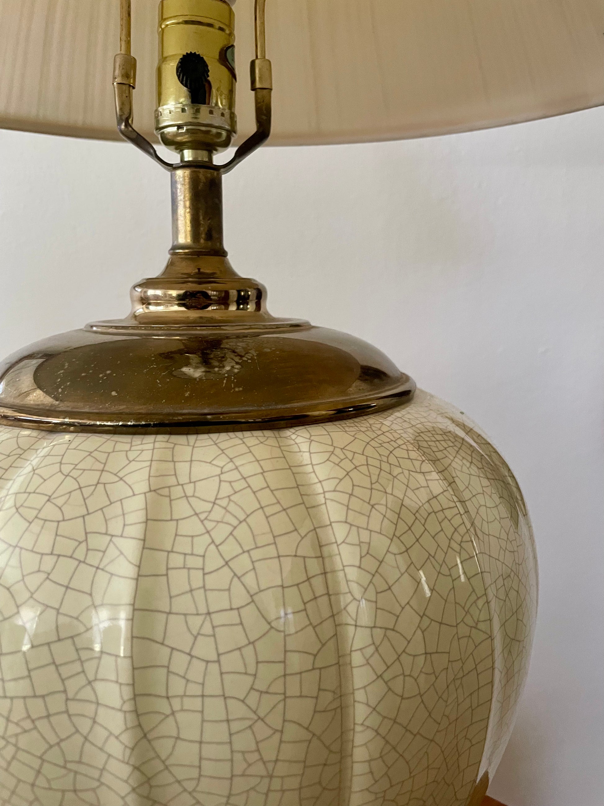 Close-up of a ceramic table lamp with a cream-colored fluted base featuring a crackle glaze pattern, a polished brass neck, and part of a beige fabric shade.