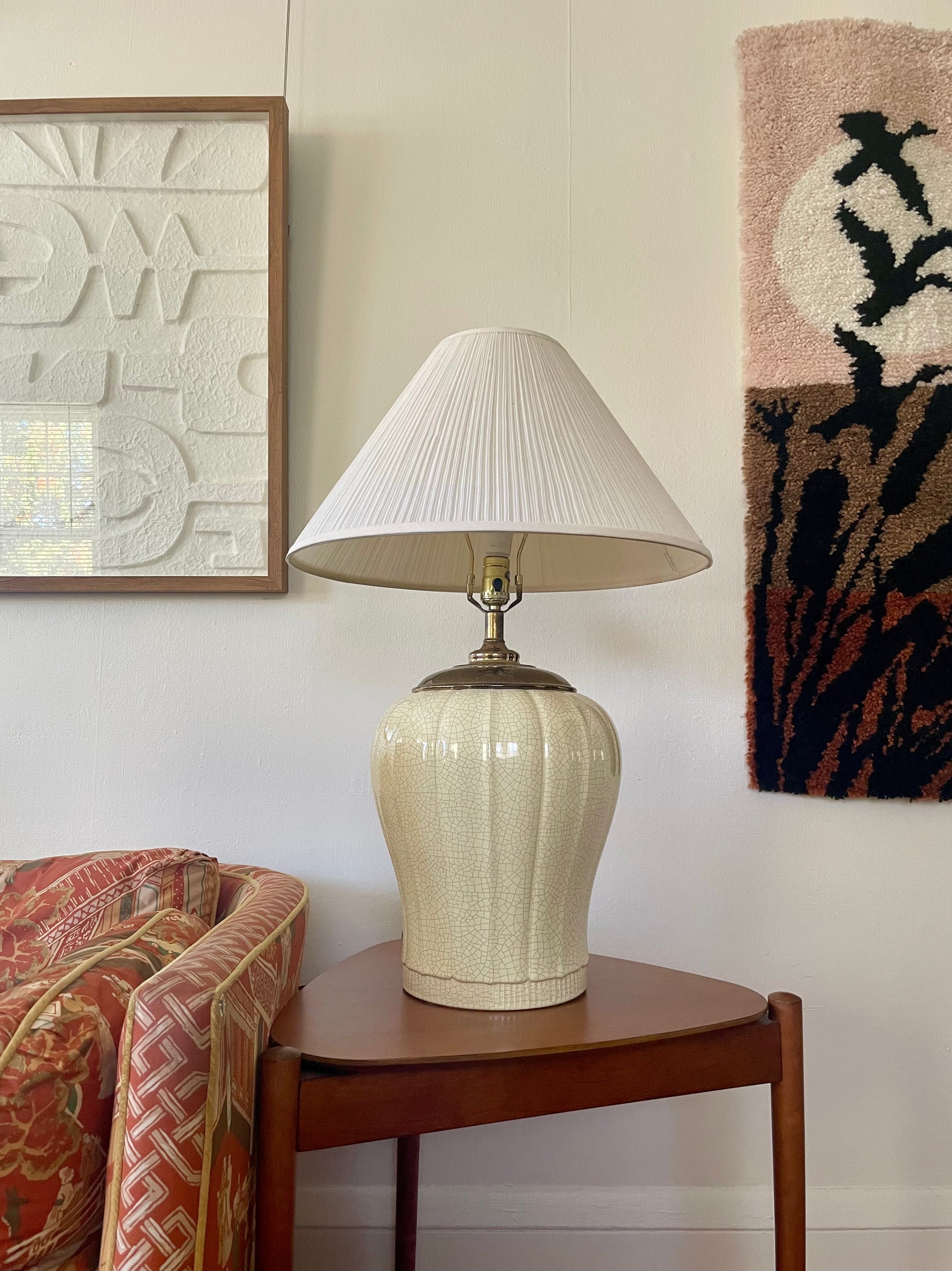 A table lamp with a ribbed cream ceramic base and a pleated white shade, styled on a wooden side table in a home interior setting.