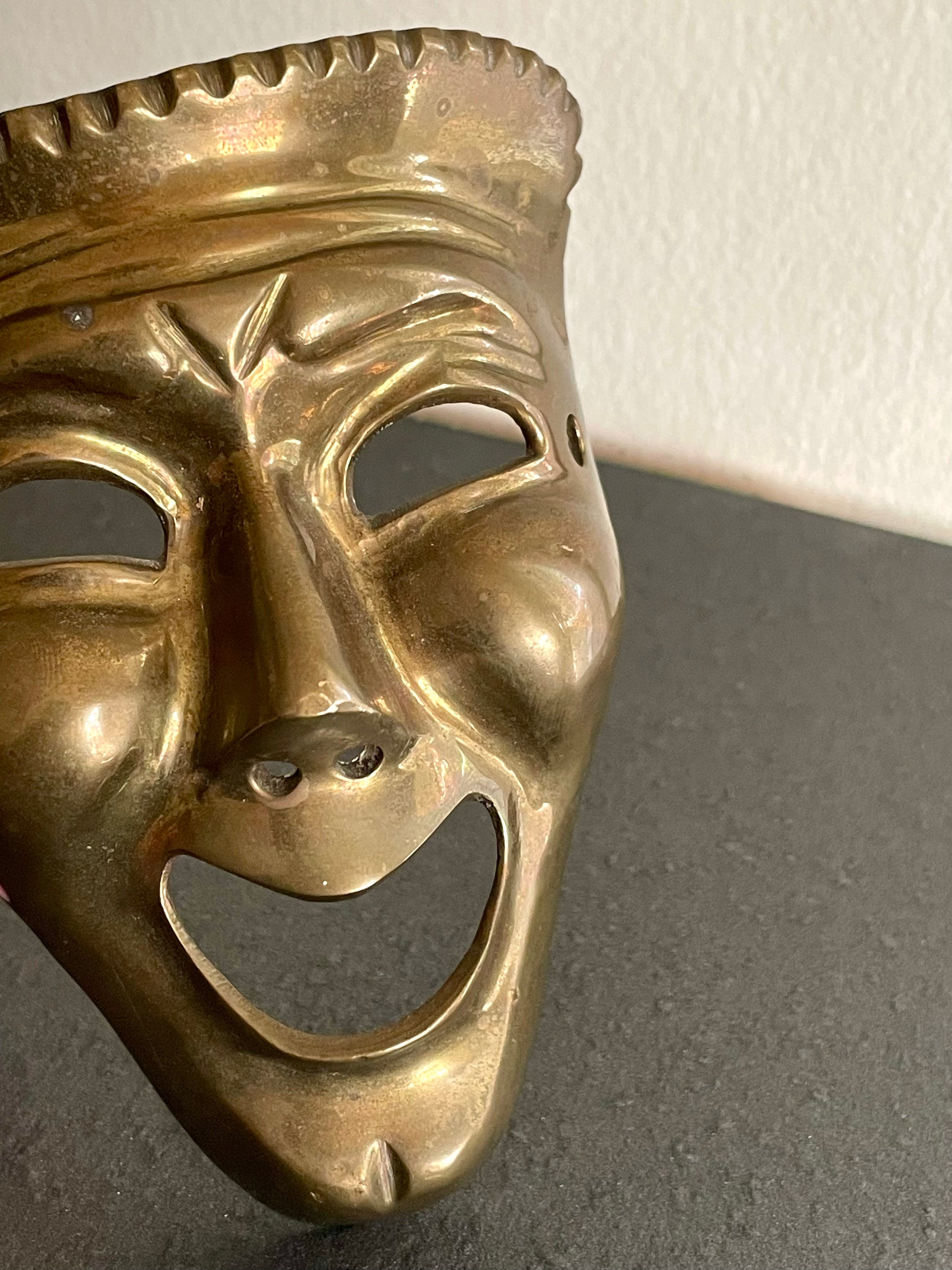 A brass theatrical comedy mask with a wide open smile, photographed close-up against a dark gray surface and a light off-white wall.