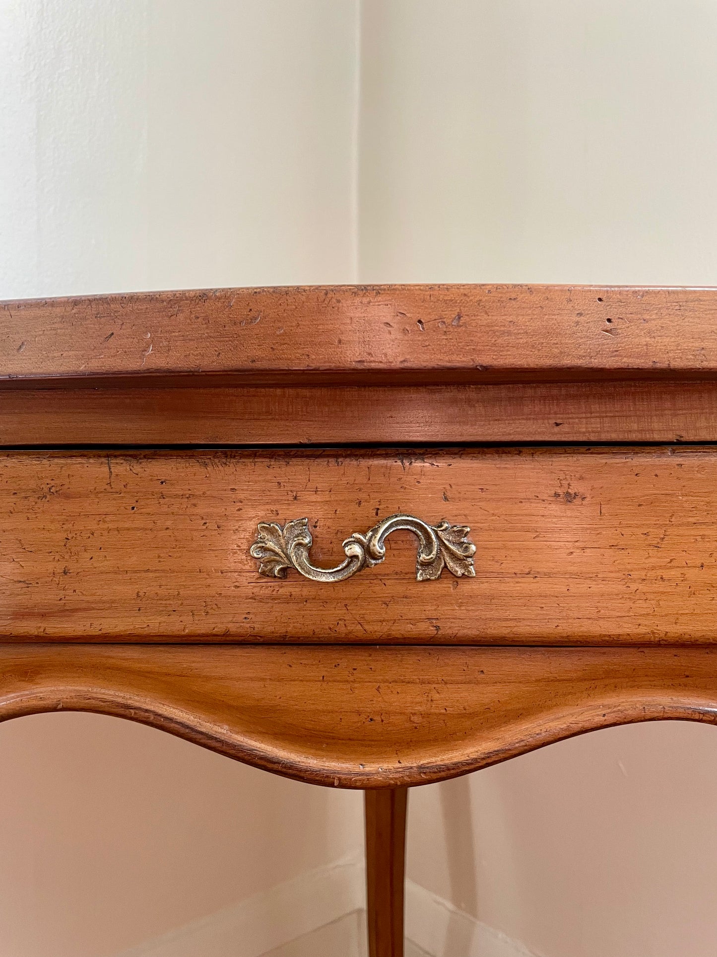 A close-up of a vintage wooden side table , showing the warm medium-brown wood grain and an ornate brass pull handle, set against a plain off-white wall corner.