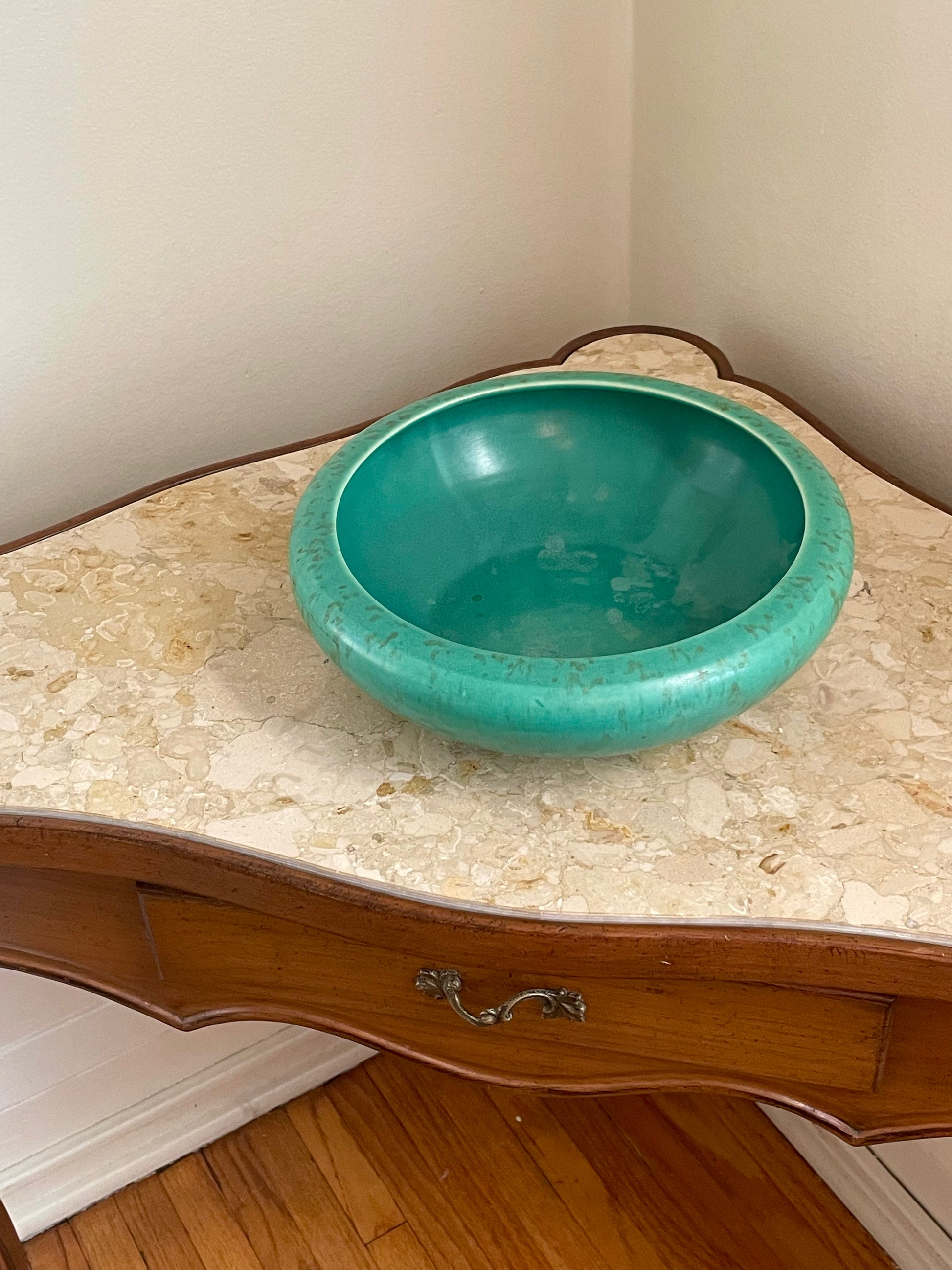 A round turquoise ceramic bowl displayed on a vintage wooden console table with a beige marble top.