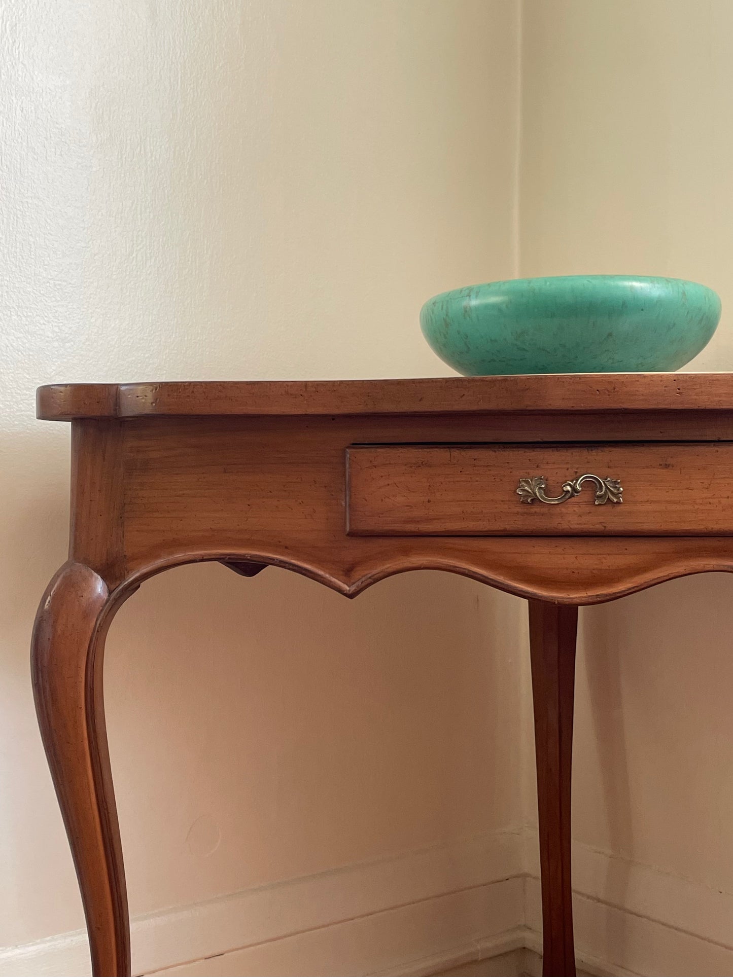 A vintage-style wooden console table with a single drawer and curved cabriole legs, shown against a cream-colored wall with a turquoise bowl on top.
