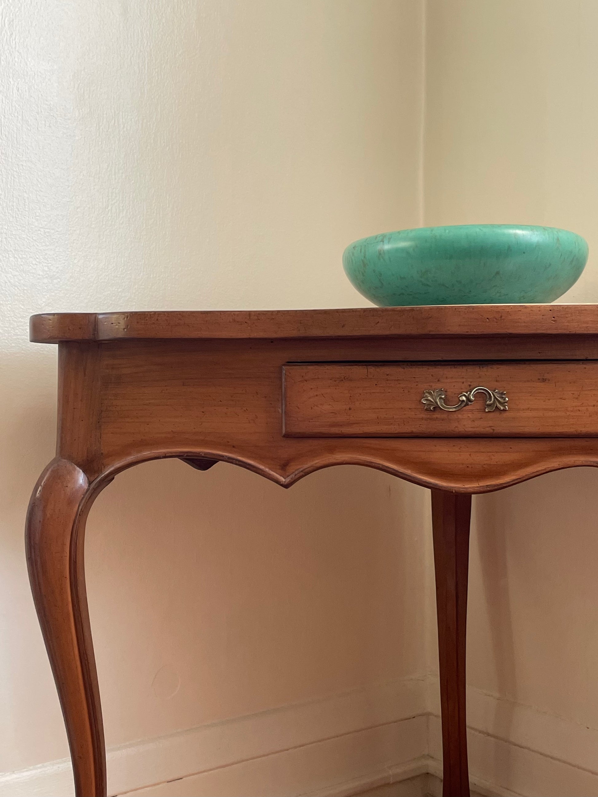 A vintage-style wooden console table with a single drawer and curved cabriole legs, shown against a cream-colored wall with a turquoise bowl on top.