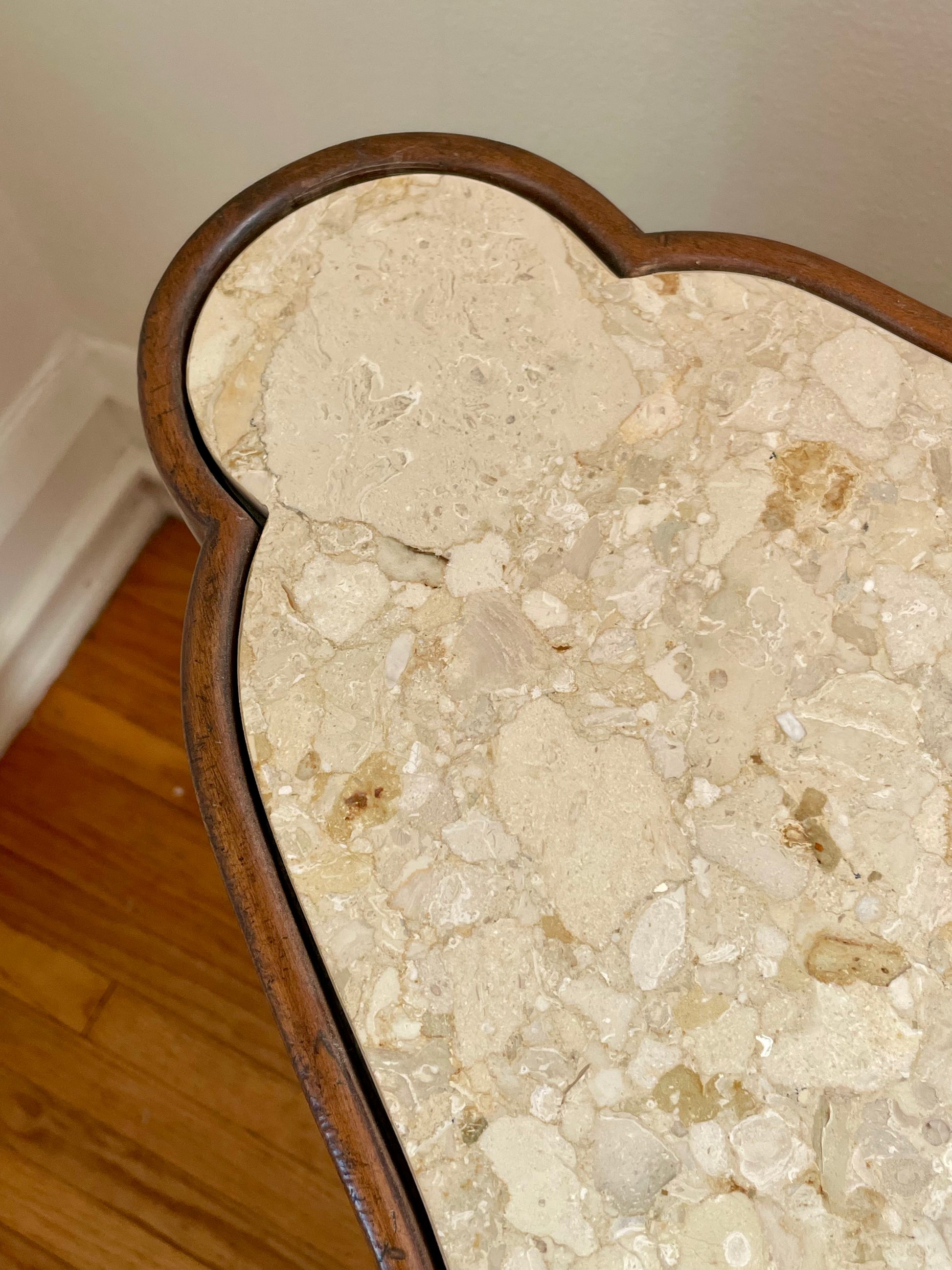 A close-up of a heart-shaped table featuring a polished beige marble stone top with natural rock fragments and a dark brown wooden frame, set against a neutral wall and hardwood floor background.