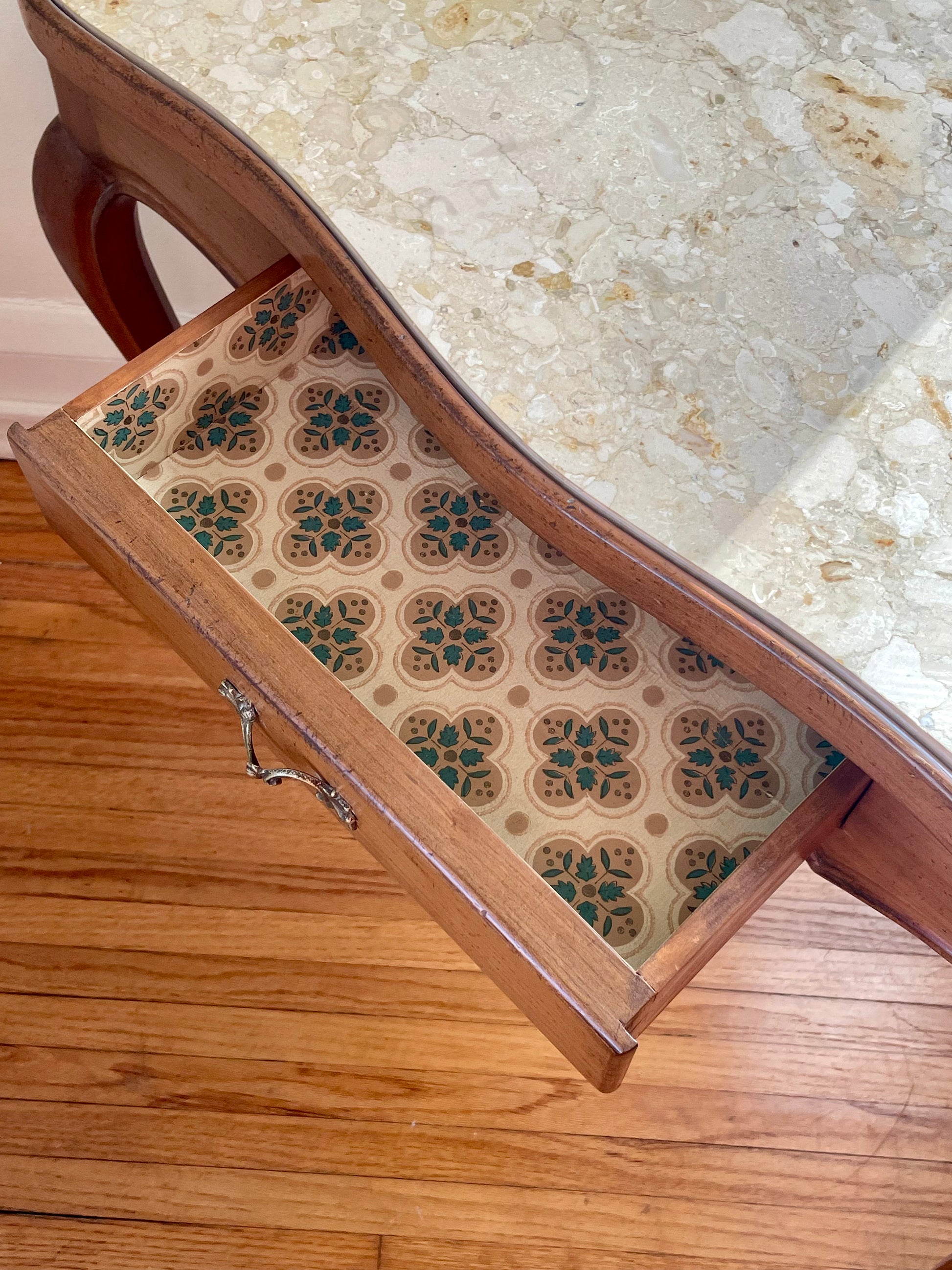 A close-up of a wooden drawer with a patterned paper lining, shown open on a hardwood floor beneath a marble-top table.