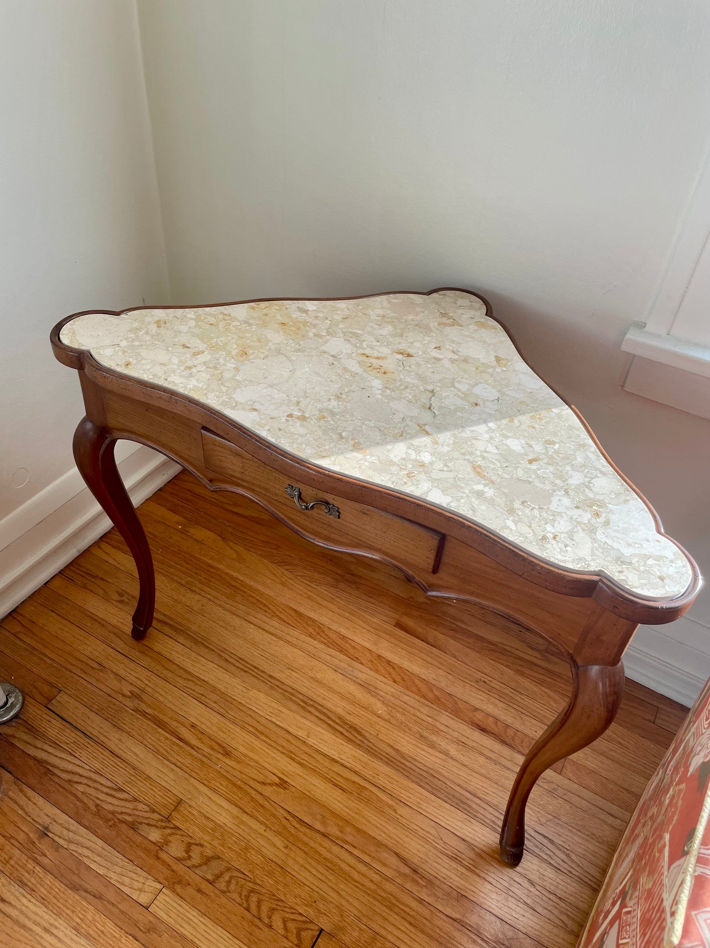 A small triangular wooden console table with a scalloped cream marble top, featuring a single drawer and curved cabriole-style legs, set on a hardwood floor against a plain wall.