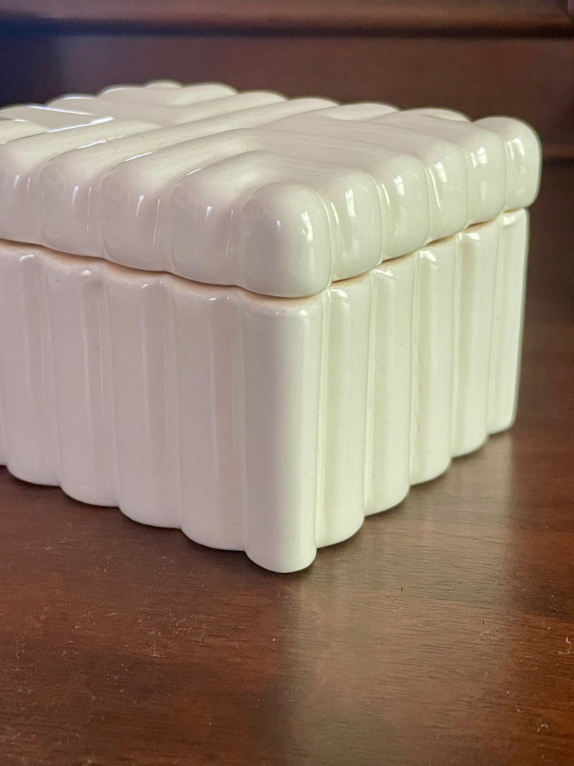 A rectangular ceramic lidded box with a glossy off-white finish, featuring a ribbed, scalloped pattern on the lid and sides, resting on a dark wooden surface.