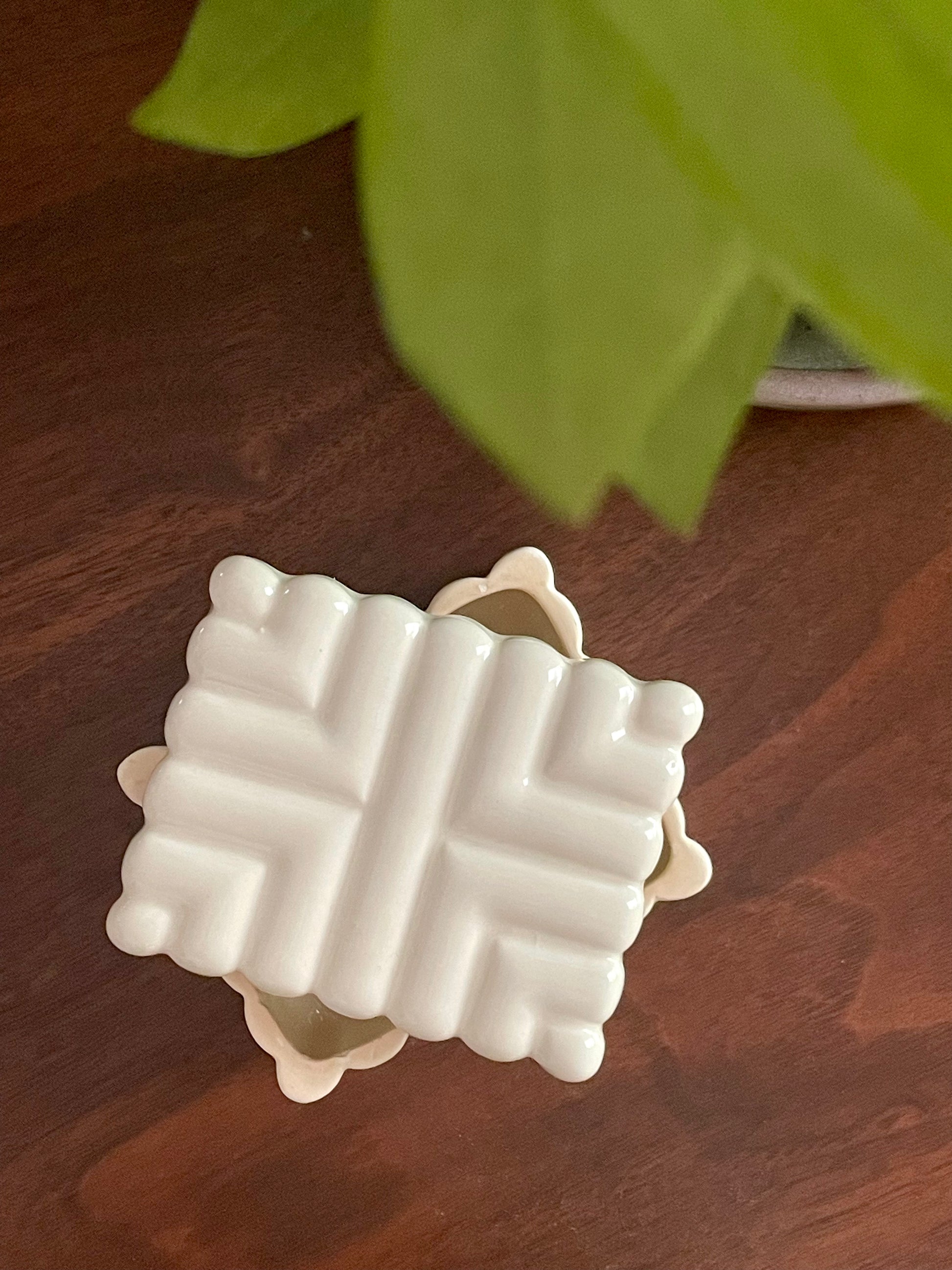 View from above of a rectangular off-white ceramic coaster with a raised geometric pattern, resting on a dark brown wooden surface with a blurred green plant leaf in the background.